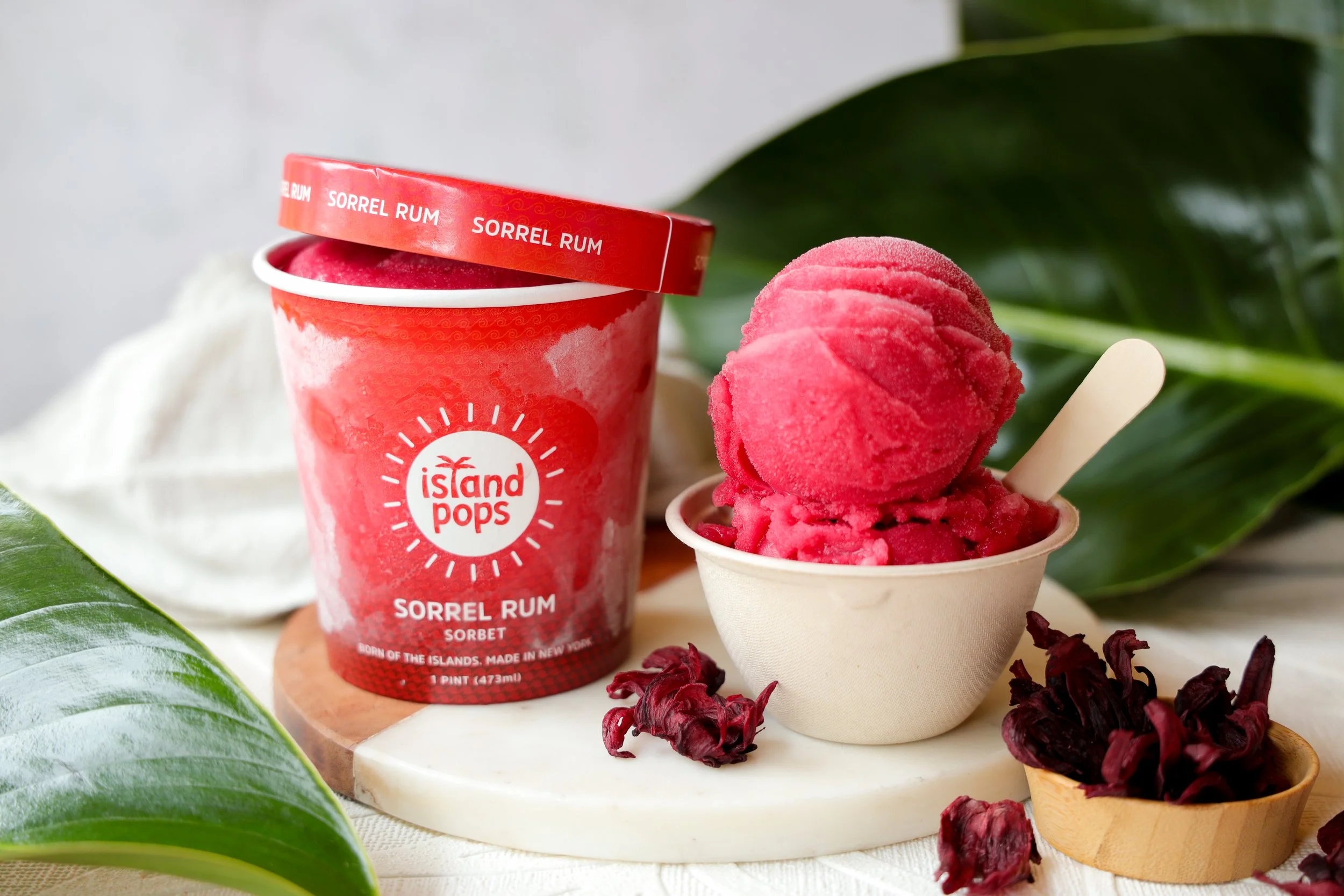 Pink sorbet ice cream in a small cup, with a wooden spoon, next to a pint container labeled 'Island Pops Sorrel Rum,' surrounded by green leaves and dried hibiscus petals.