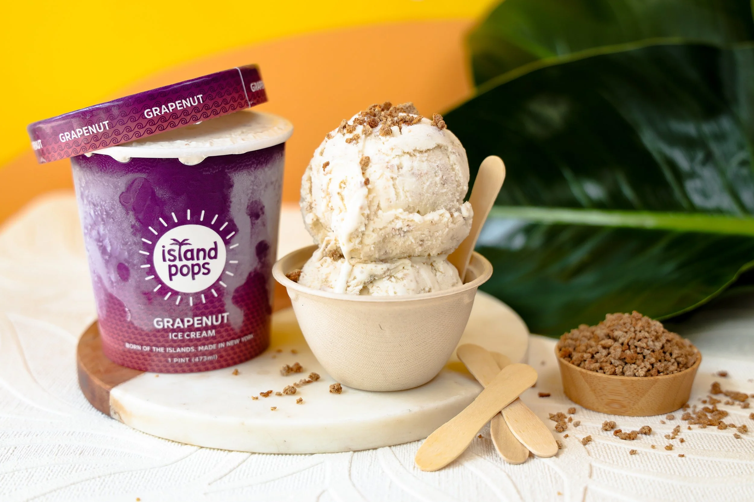 Grapenut ice cream from Island Pops, with two scoops in a beige cup topped with crumbles, alongside a partially opened container of Grapenut ice cream and two small wooden spoons, on a white textured surface with green leaves in the background.