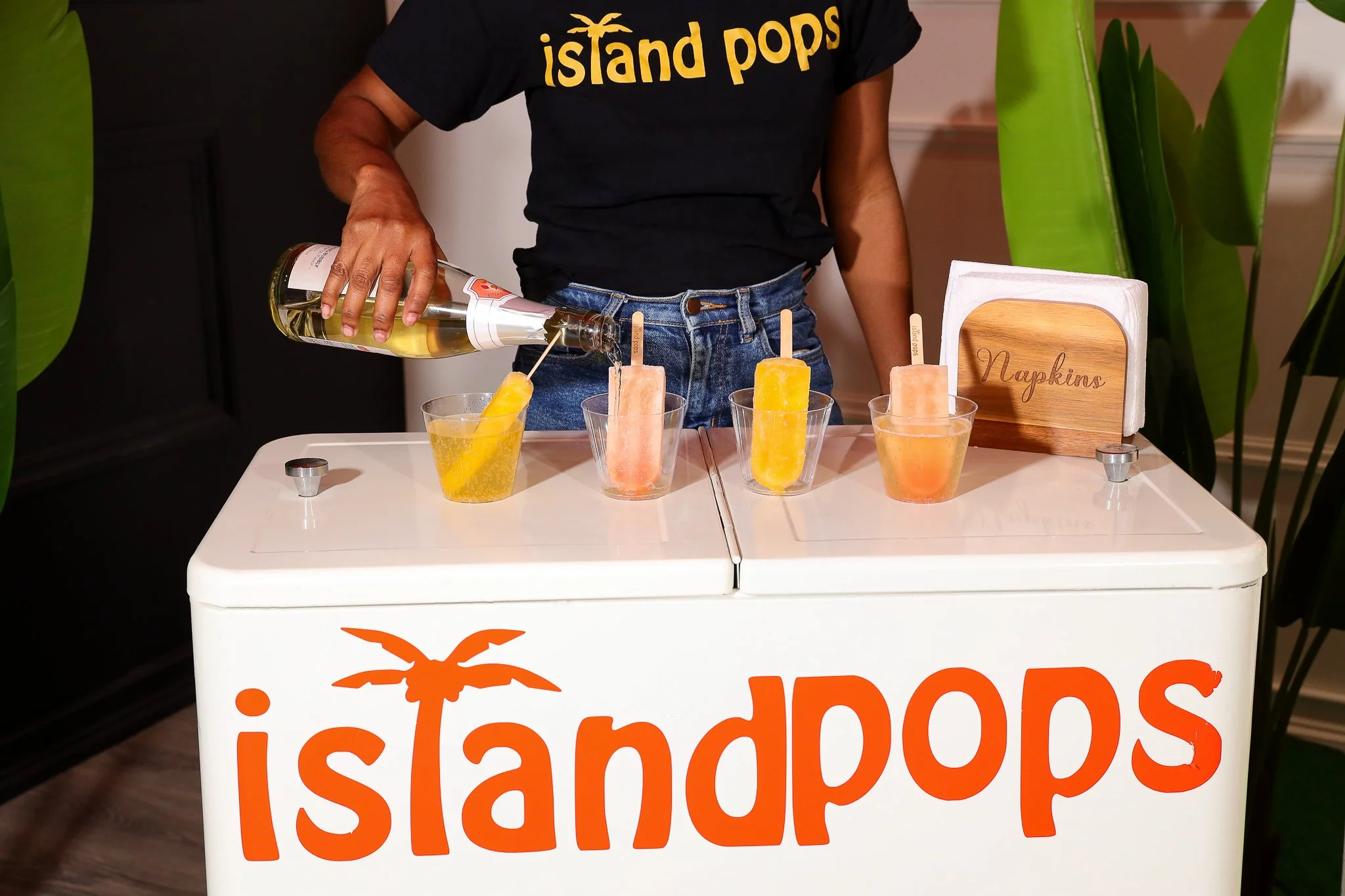 Person pouring a yellow drink into a clear cup at an island pops stand with three colorful popsicles on sticks displayed.