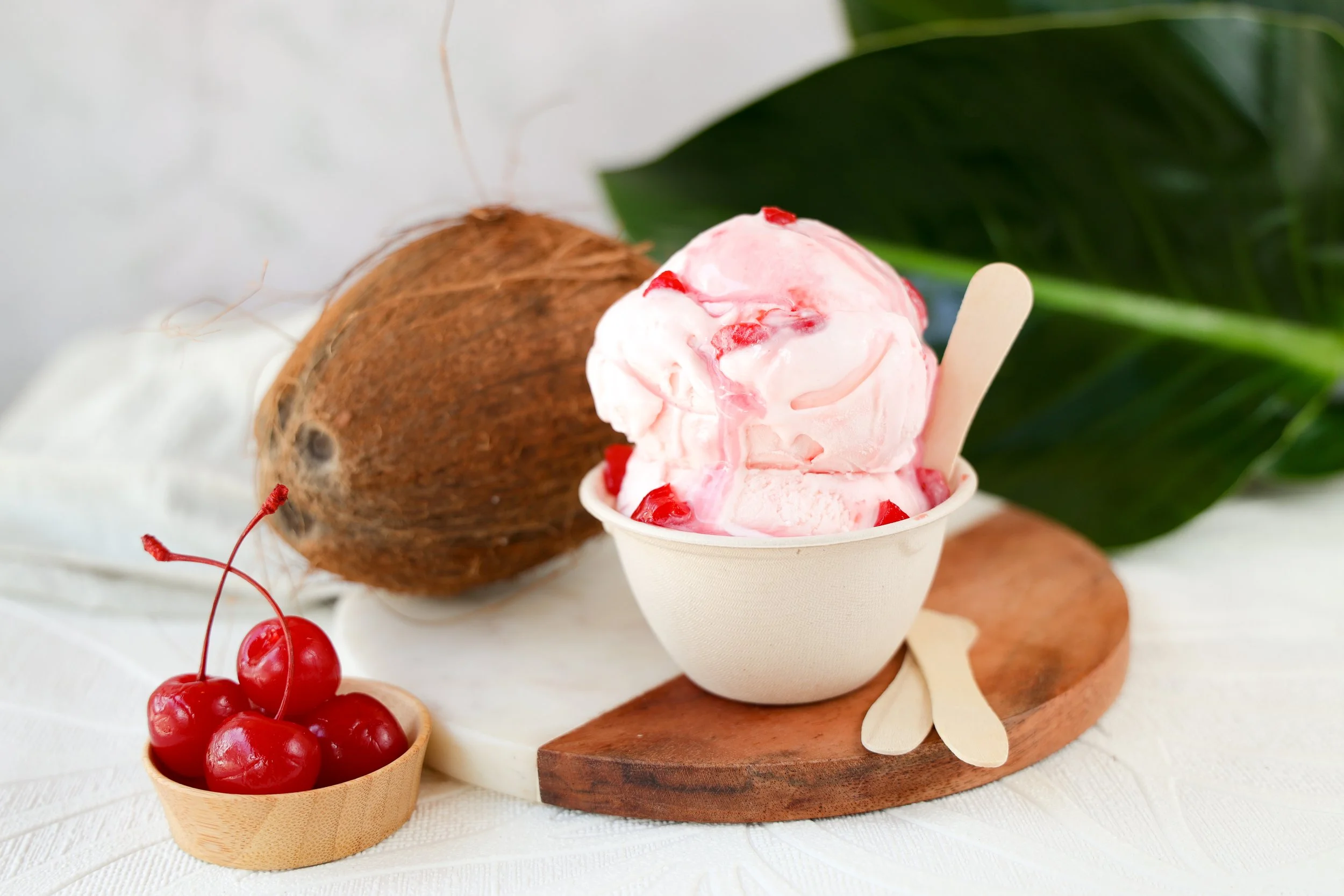 A bowl of cherry-flavored ice cream with cherry pieces served in a white cup, with a wooden spoon. Fresh cherries in a small bamboo bowl and coconuts are in the background, along with green leaves, all arranged on a white surface with a wooden board