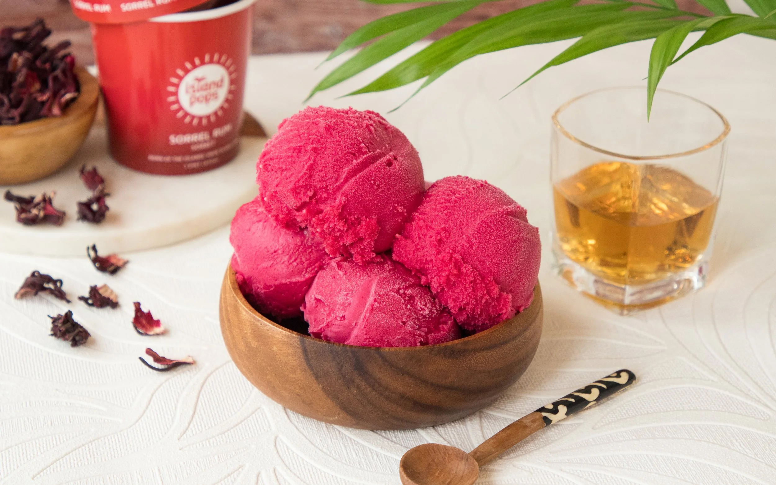 Bowl of three scoops of sorrel rum sorbet on a white surface, with a glass of whiskey, a wooden spoon, and a container of sorbet in the background.