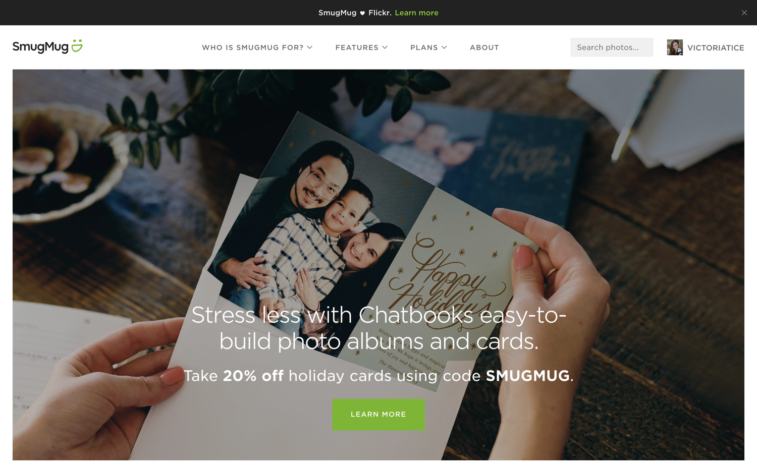 Close-up of a person holding a photo album with family photos and holiday cards, on a wooden table with greenery and paper in the background.