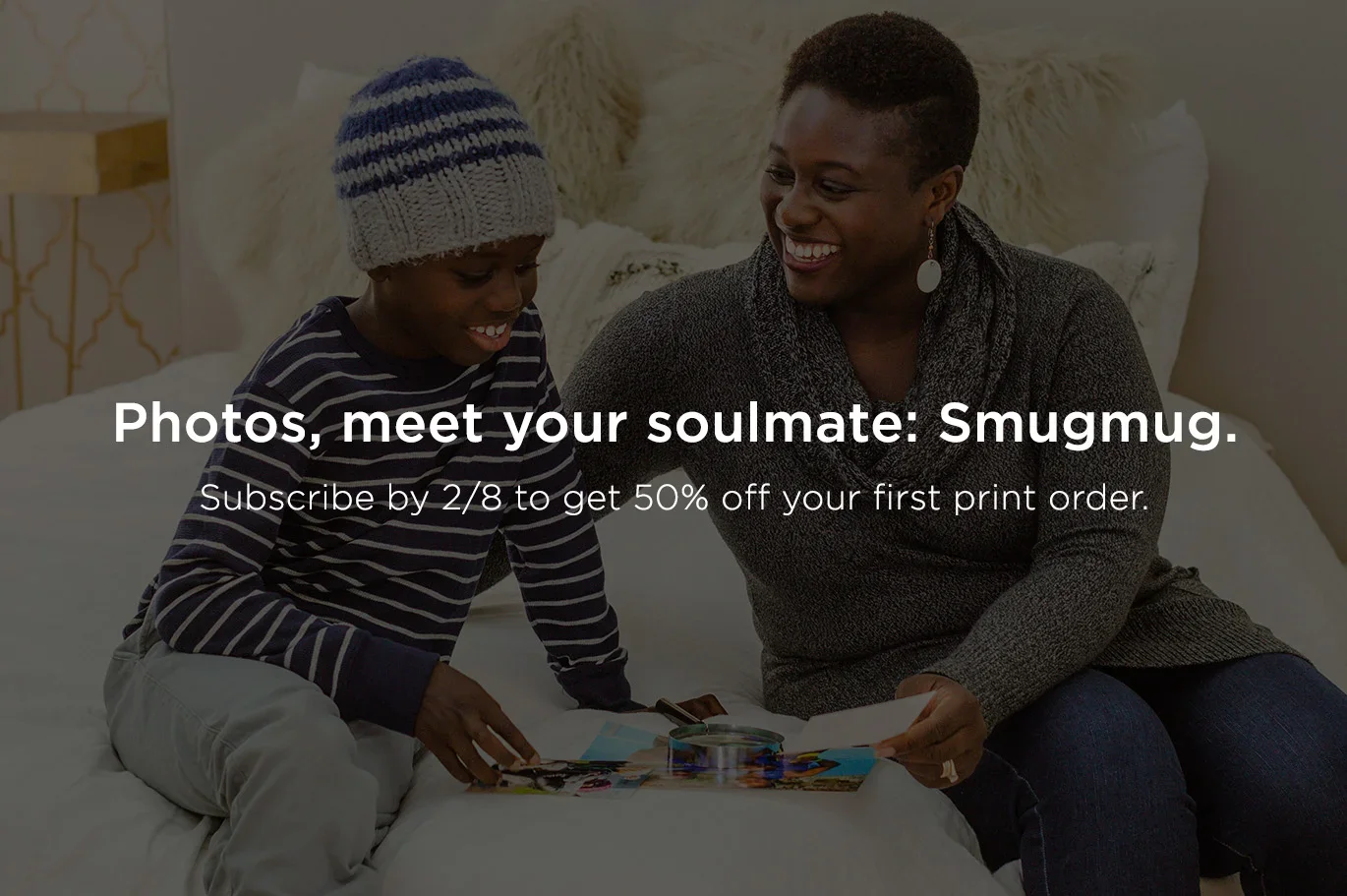 A woman and a young boy sitting on a bed, smiling and looking at photos. The boy is wearing a striped sweater and a knit hat. The woman is wearing a dark gray sweater and earrings.