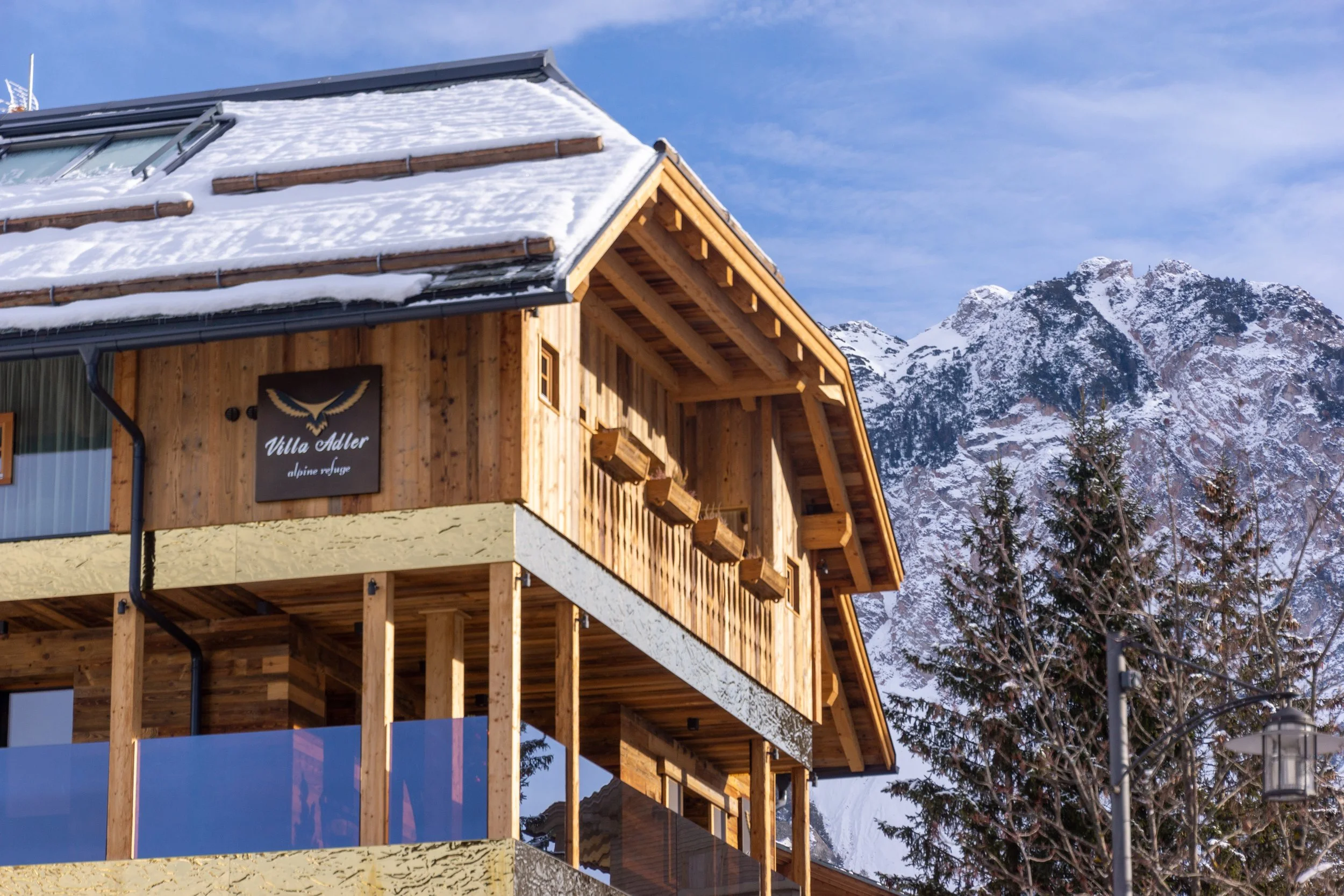 Edificio in stile chalet di legno con vista sugli alti Monti delle Alpi, parzialmente innevati, sotto un cielo blu con alcune nuvole.