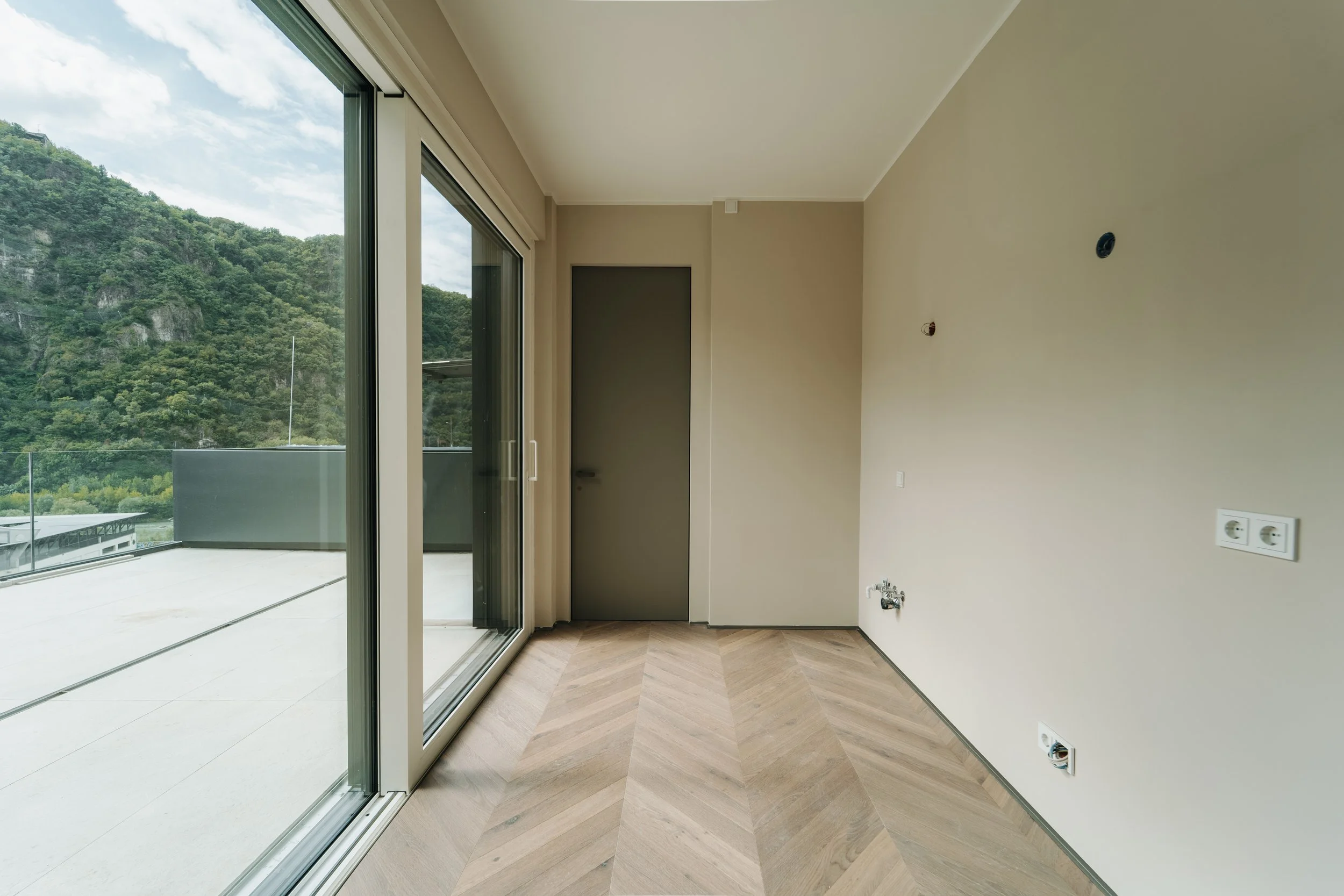 Stanza vuota con pareti beige, pavimento a motivo a spina di pesce, grande finestra scorrevole che dà su un balcone con vista su montagne verdi, porta chiusa e prese elettriche sulla parete.