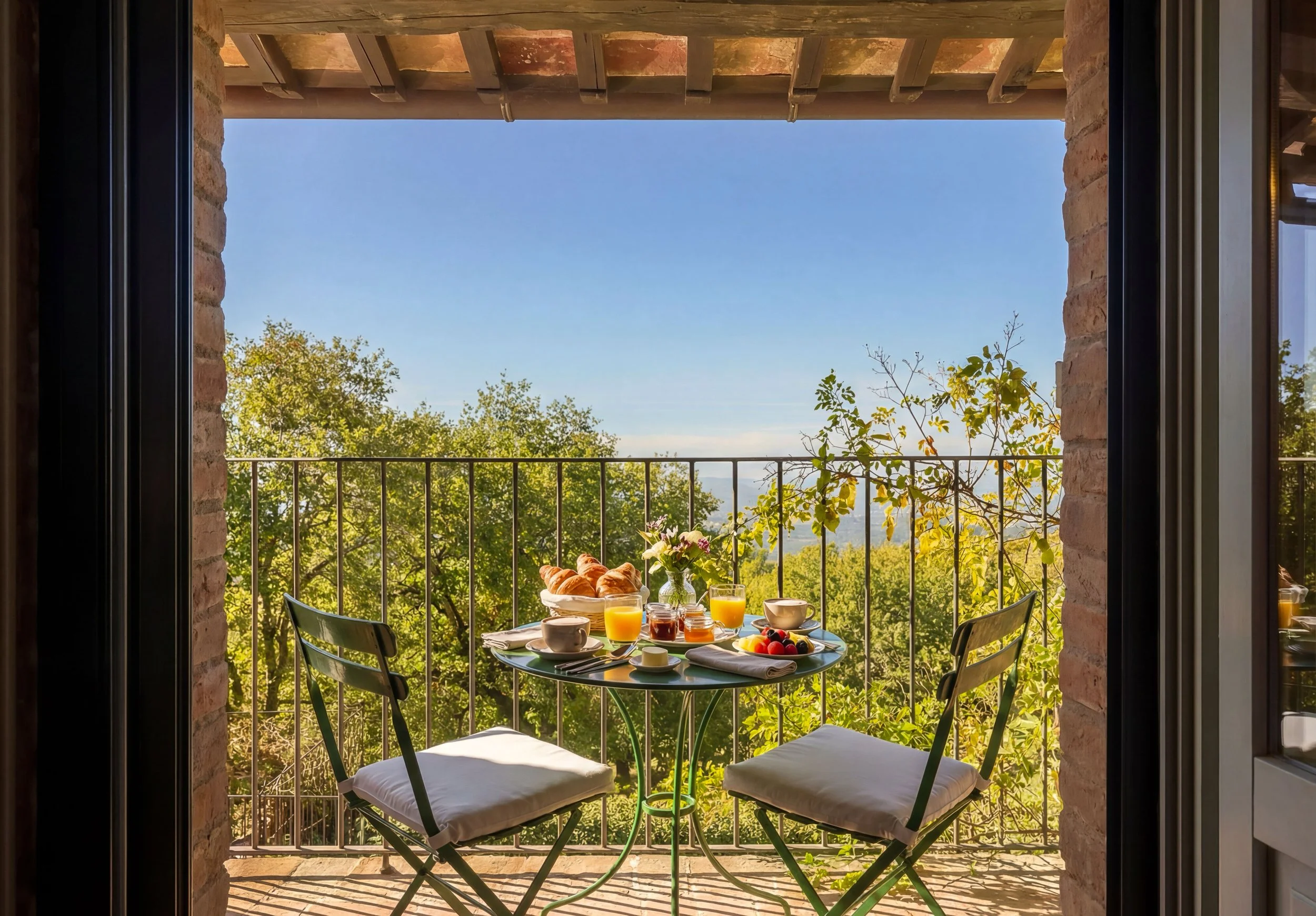 Colazione su un balcone con vista su alberi e cielo azzurro, tavolo con croissant, succhi, tazze e frutta, due sedie con cuscini.