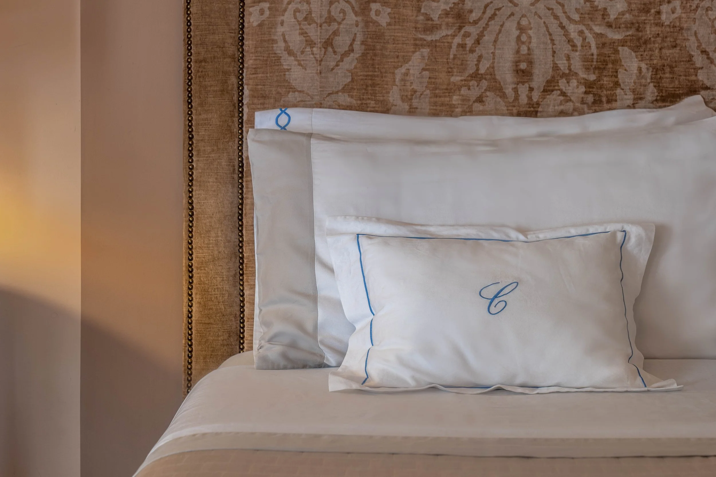 Letto con due cuscini bianchi con cuciture blu e lettera C cucita sul cuscino più piccolo, e testiera imbottita con motivo floreale in tessuto marrone.