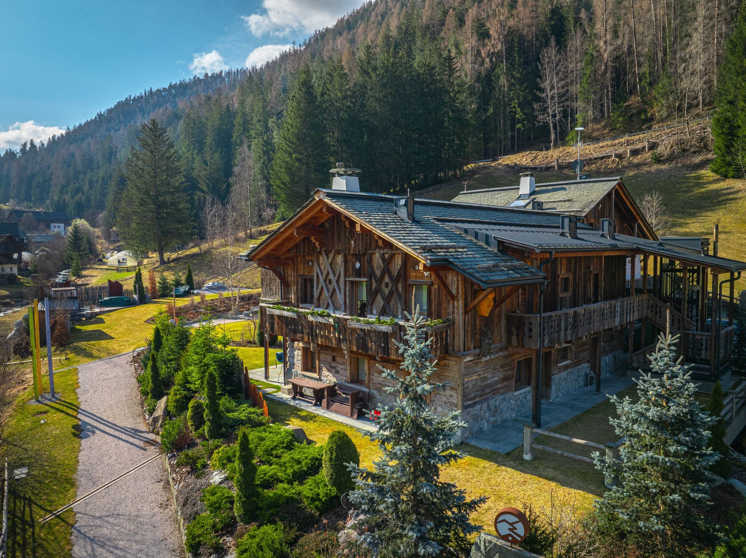 Servizio fotografico chalet  in legno in Alto Adige a San Vigilio di Marebbe, terrazza in legno circondata da alberi e un giardino ben curato, con un percorso di ghiaia, ai piedi di una catena montuosa.