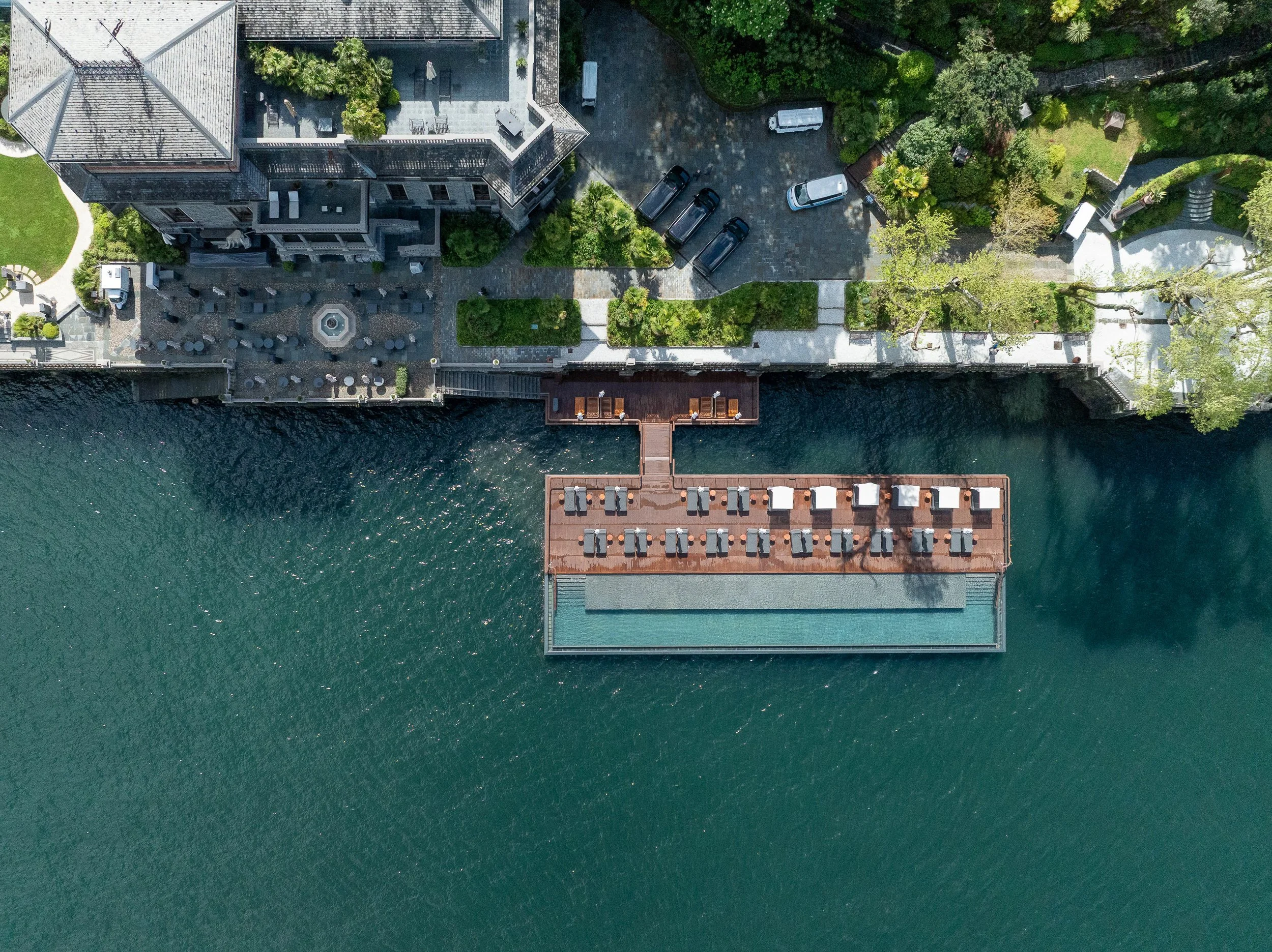 Veduta aerea di un edificio di grandi dimensioni vicino a un corpo d'acqua, con un'area di relax sul lago dotata di lettini e ombrelloni, e un incrocio con parcheggio e giardini.