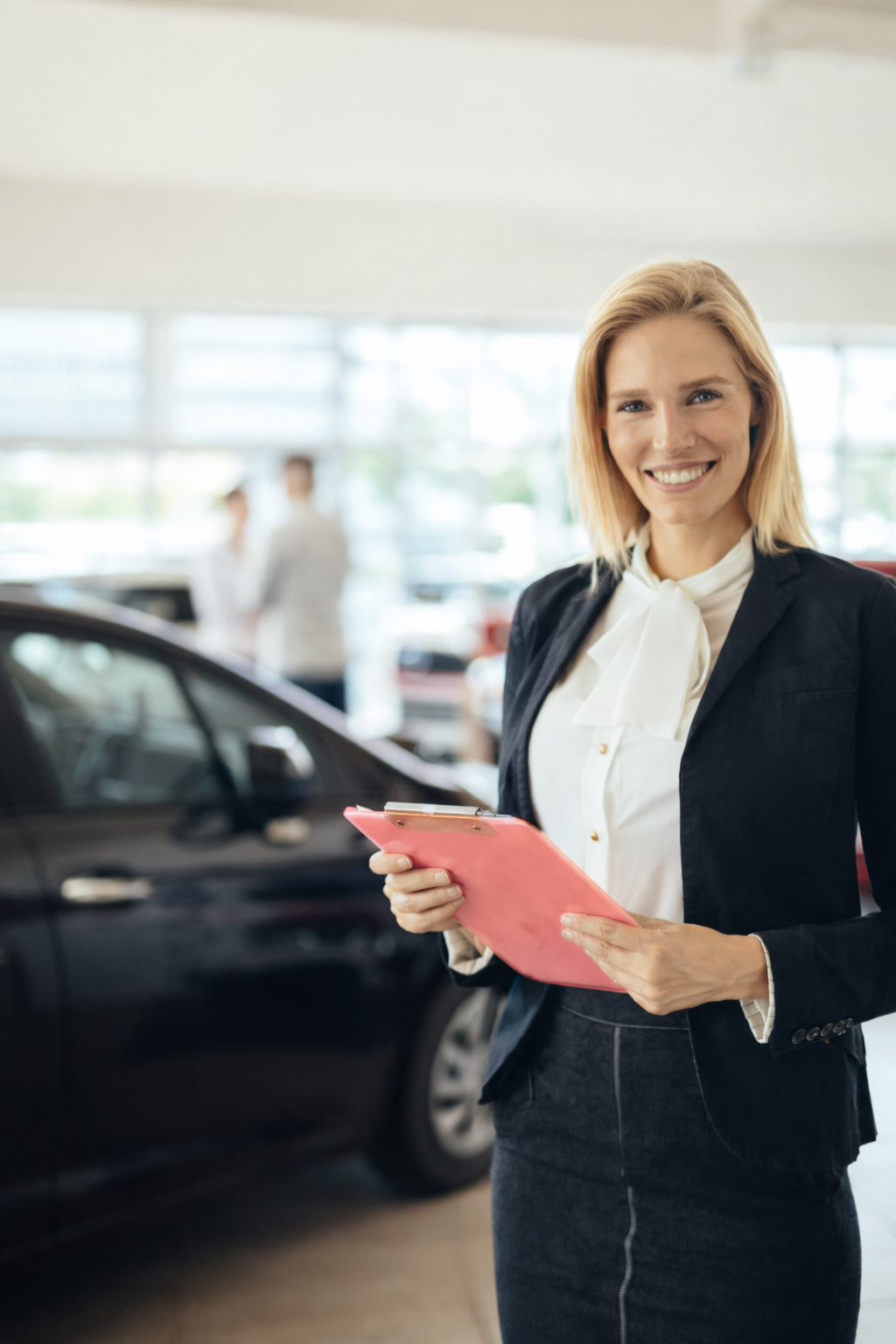 Eine lächelnde Frau in Geschäftskleidung hält einen rosa Klemmbrett und steht vor einem schwarzen Auto in einem Autohaus.
