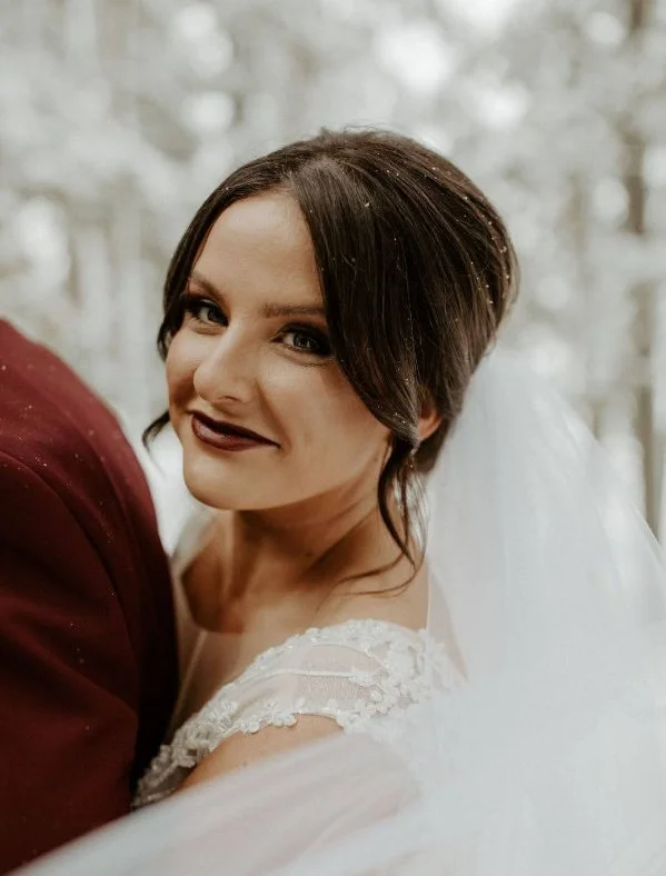Wedding bride with short dark hair and dark lipstick smiling outdoors with blurred trees in the background, wearing a lace wedding dress and white veil.
