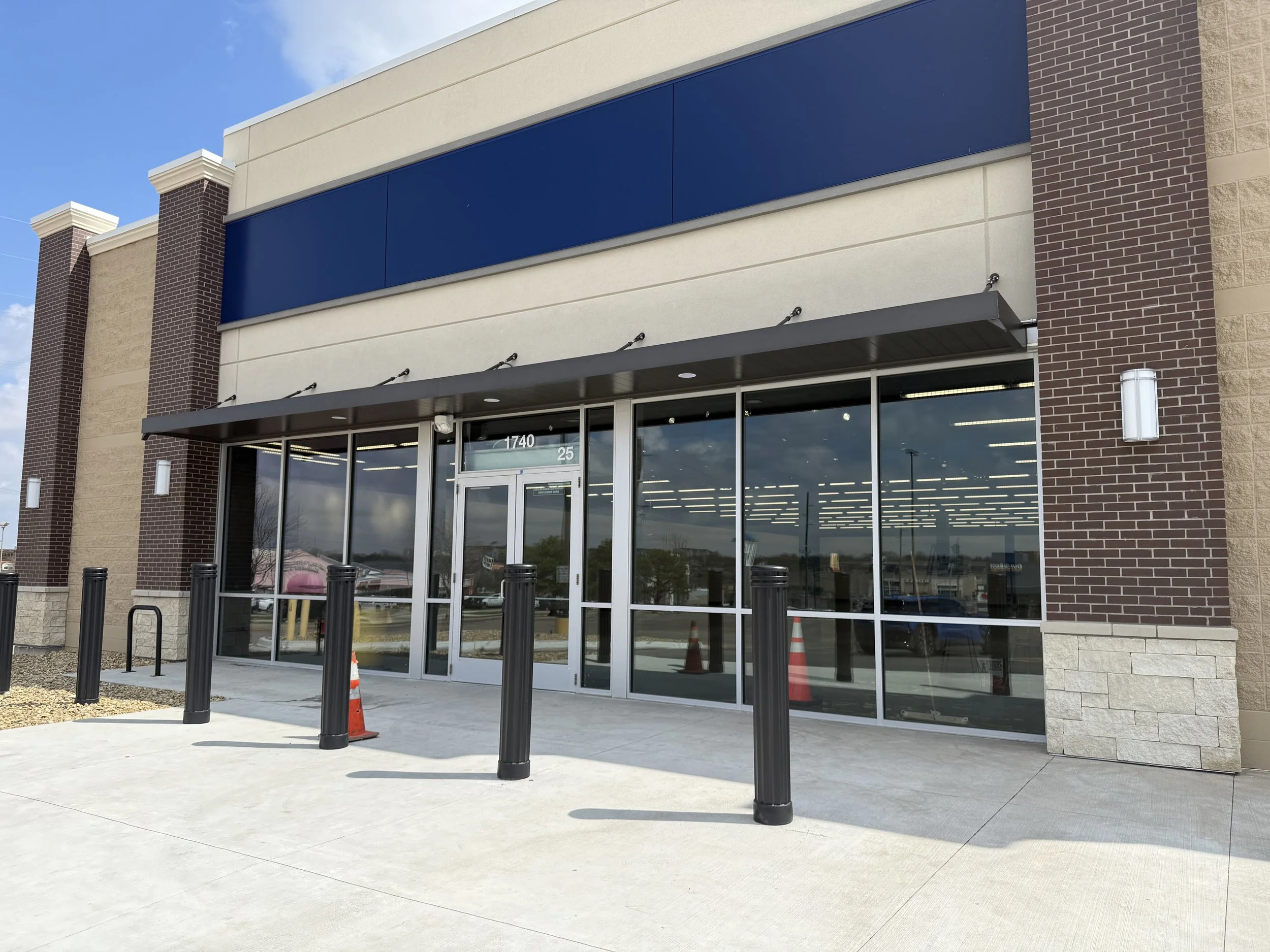New Sketchers store awning installed. Front exterior view of a new retail store with large glass windows, a glass door, and black bollards, with a blue sign space above the entrance, and a small traffic cone on the sidewalk in front.