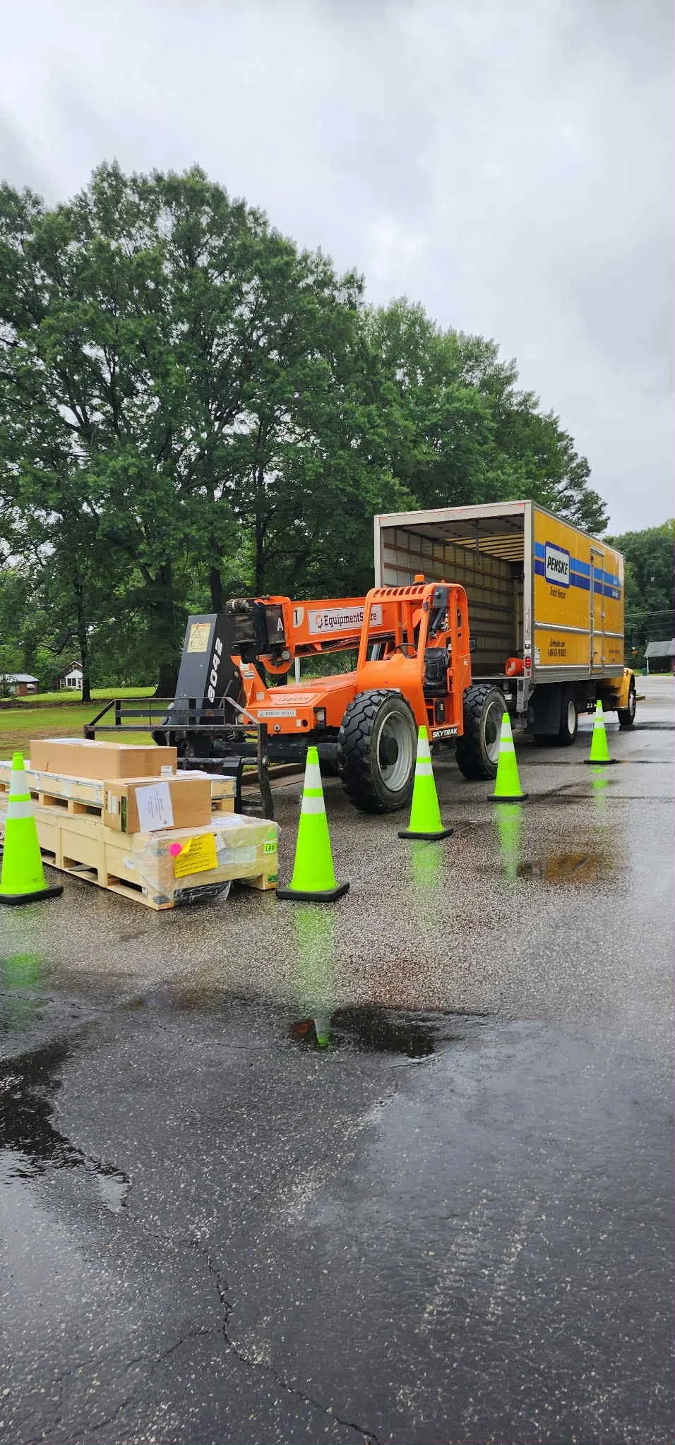 A delivery truck parked on a wet road with green traffic cones around it, a pallet with boxes in front, and trees in the background.