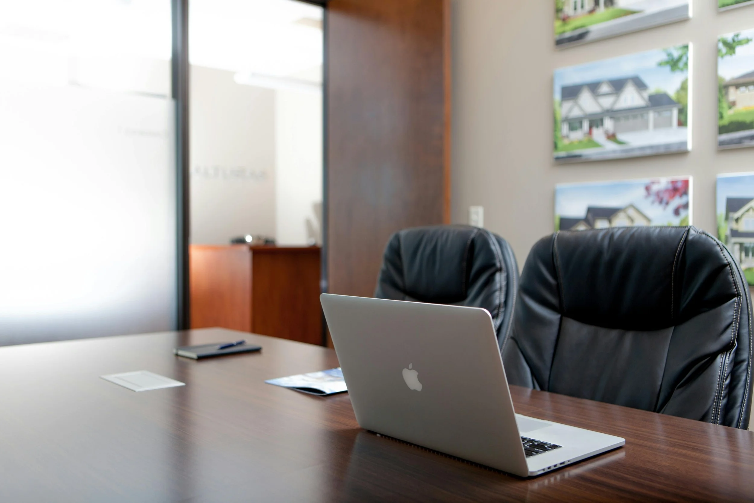An empty conference room with a wooden table, two black leather chairs, and a silver Apple MacBook laptop on the table. There are some papers and a pen on the table, with framed house images on the wall and a frosted glass door in the background.