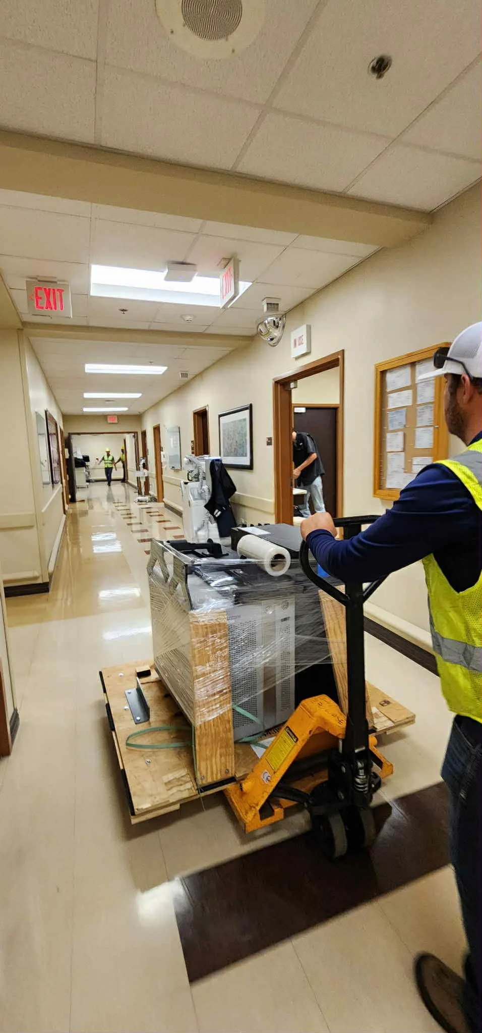 A worker operating a pallet jack with a large electronic device wrapped in plastic in a beige corridor, with another worker in the distance.