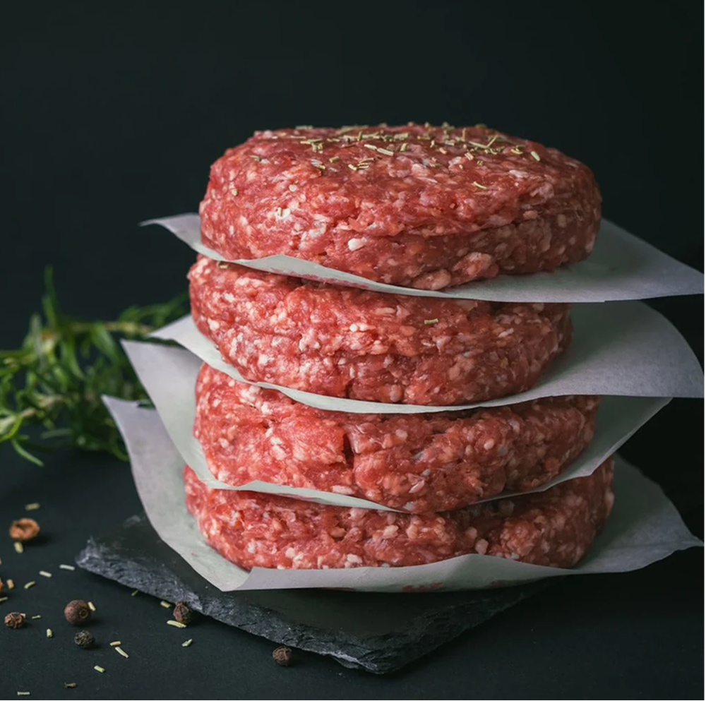 Stack of four raw beef hamburger patties with herbs on top, separated by parchment paper, on a black slate board with some peppercorns and herbs around.