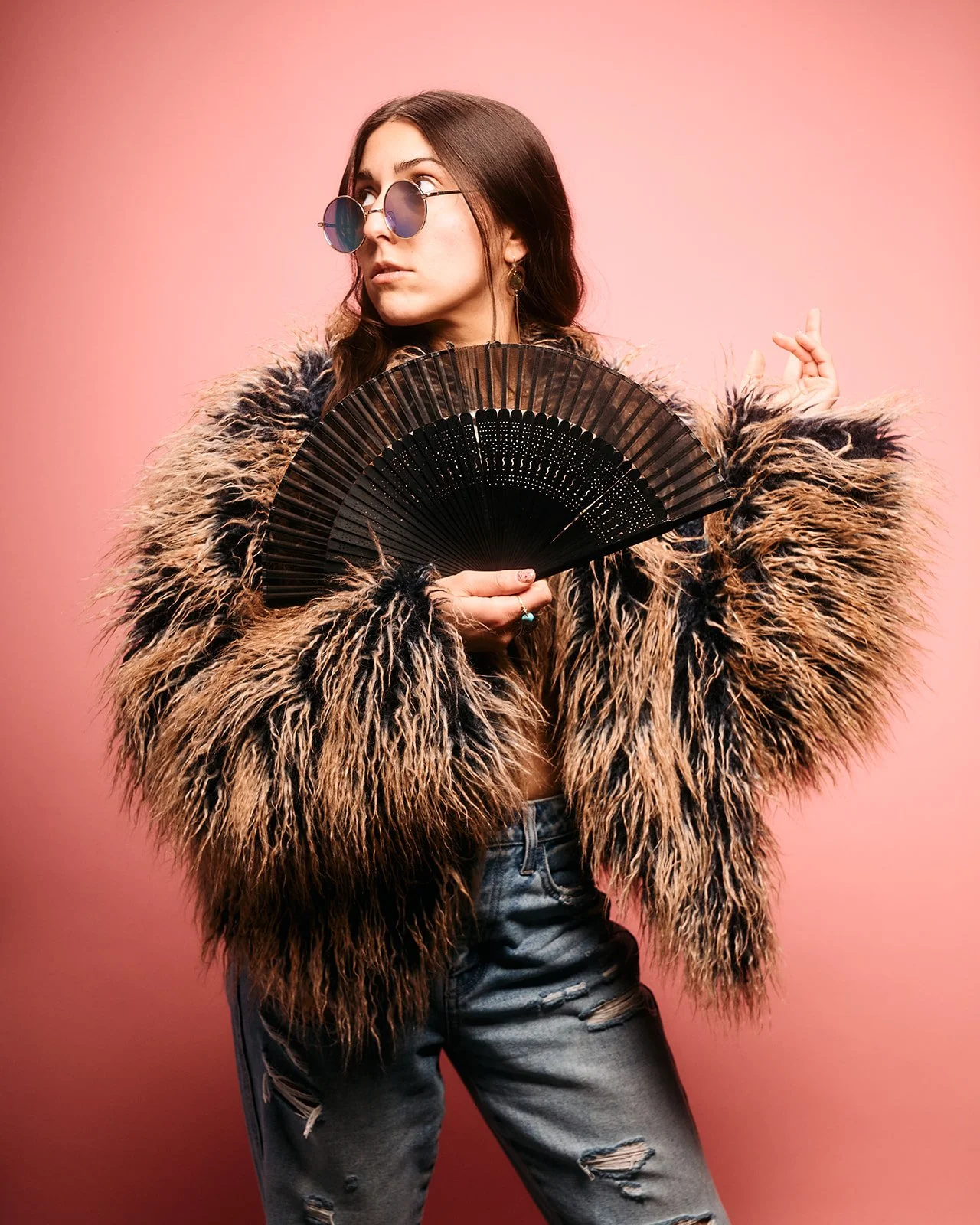 Artist headshot in faux fur coat, sunglasses, jeans & holding a fan. DJ, Healer, Yoga Teacher, Reiki Master, Vocalist and Songwriter.