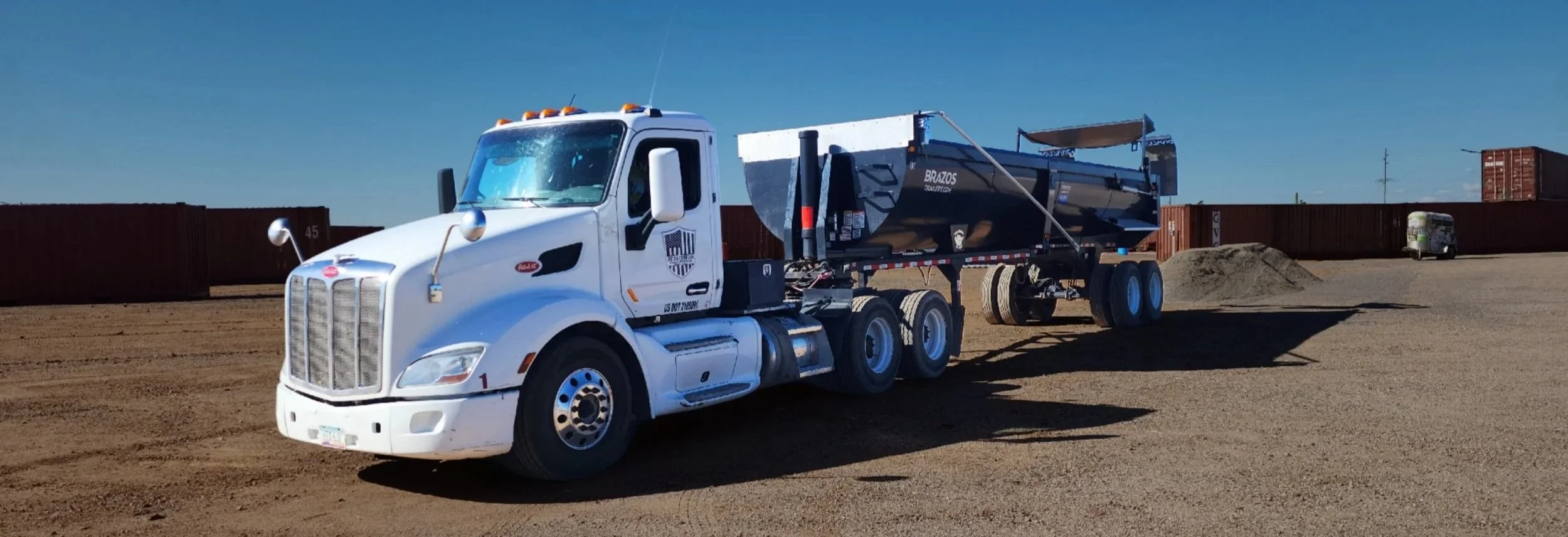 A white Peterbilt semi-truck with a black dump trailer parked on a dirt lot under a clear blue sky.