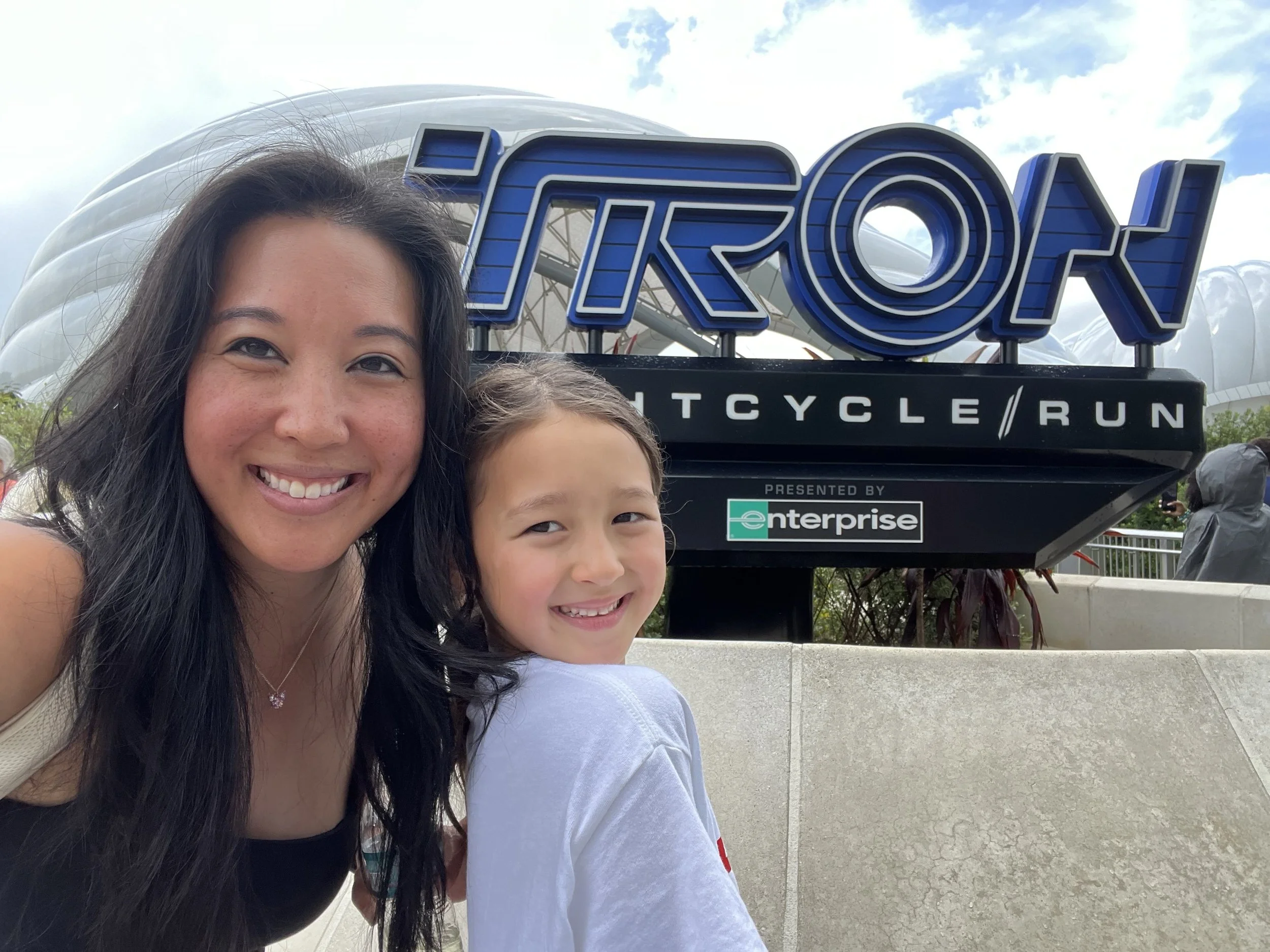A woman and a young girl smiling for a selfie in front of the Tron Lightcycle/Run sign at a theme park, with cloudy skies in the background.