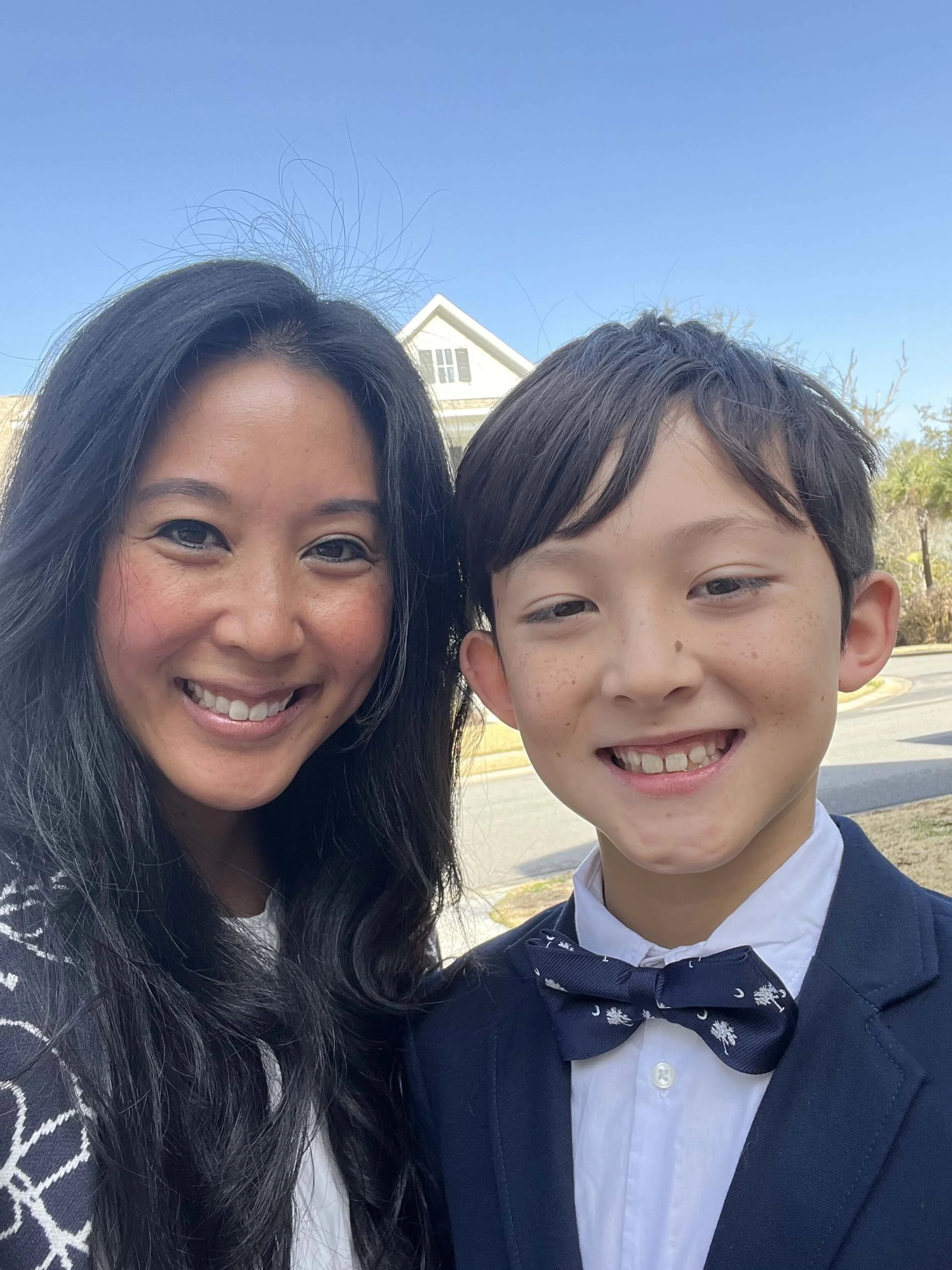 A smiling woman with long dark hair and a young boy with short dark hair, wearing a tuxedo and bow tie, taking a selfie outdoors on a sunny day.