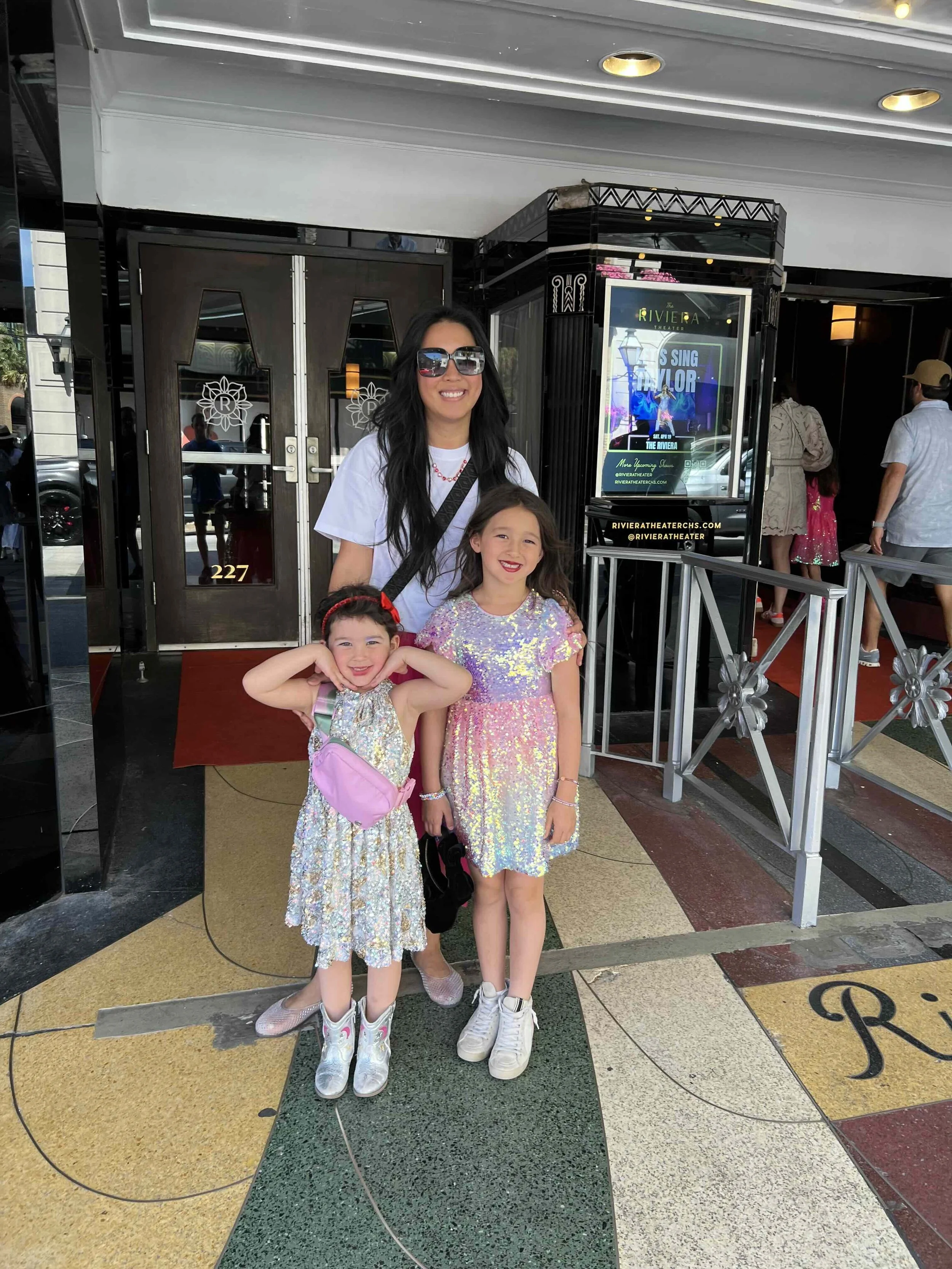 Three females standing together outside a theater. The adult woman is smiling, wearing sunglasses, a white t-shirt, and carrying a black shoulder bag. The two young girls in front are smiling; one has her arms raised, wearing a sequin dress and a pin