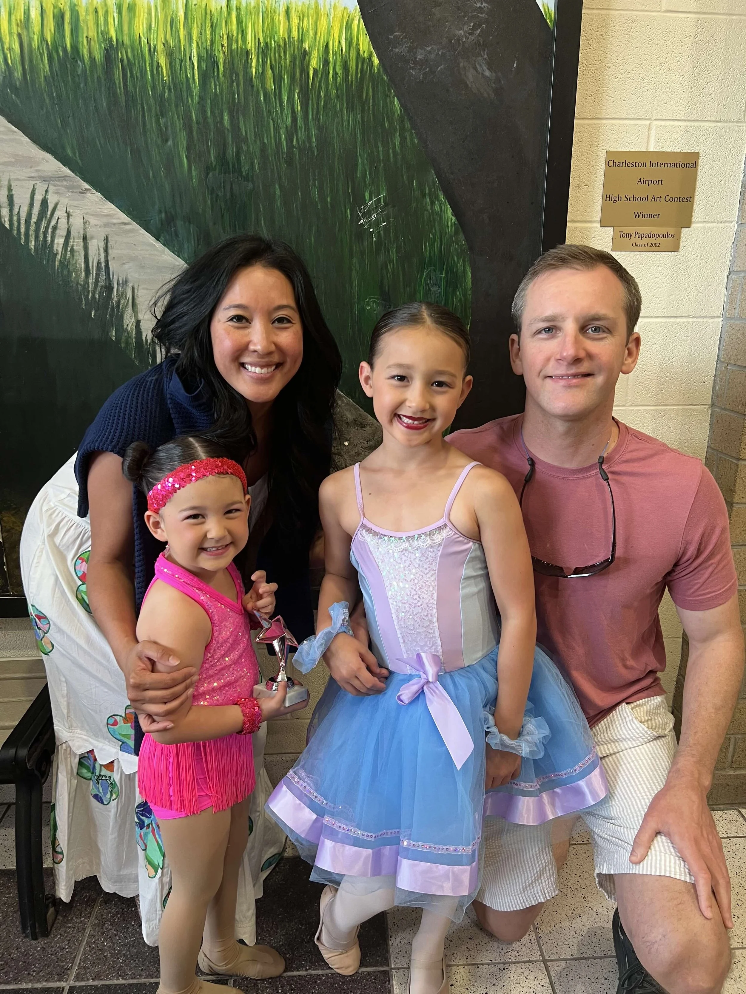 A family of five posing together indoors, with two young girls dressed in colorful costumes, one in a pink dress and the other in a blue dress, and two adults, a woman with long black hair and a man with short light brown hair, smiling. The backgroun