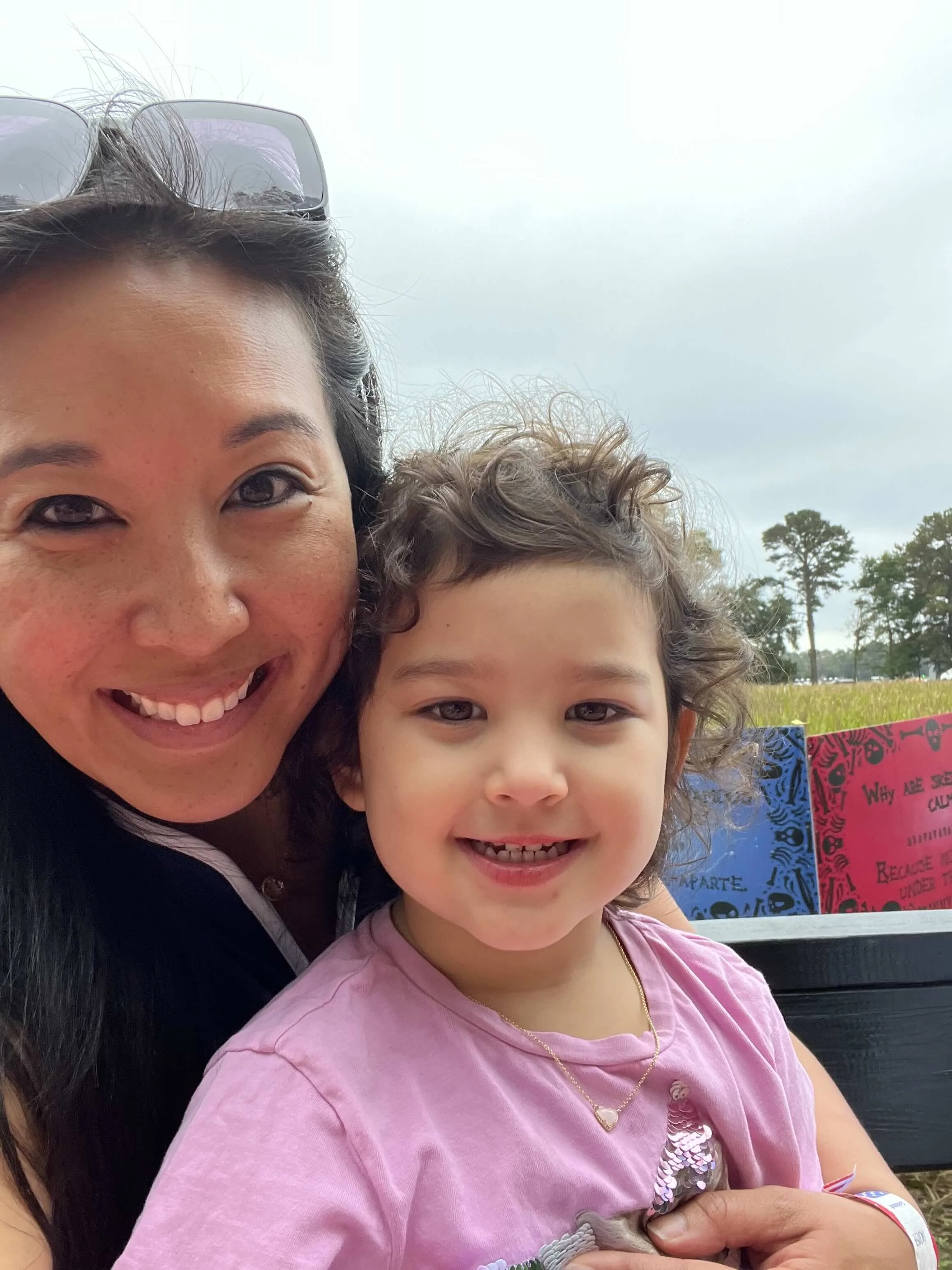 A woman and a young girl taking a selfie outdoors with trees and sky in the background.