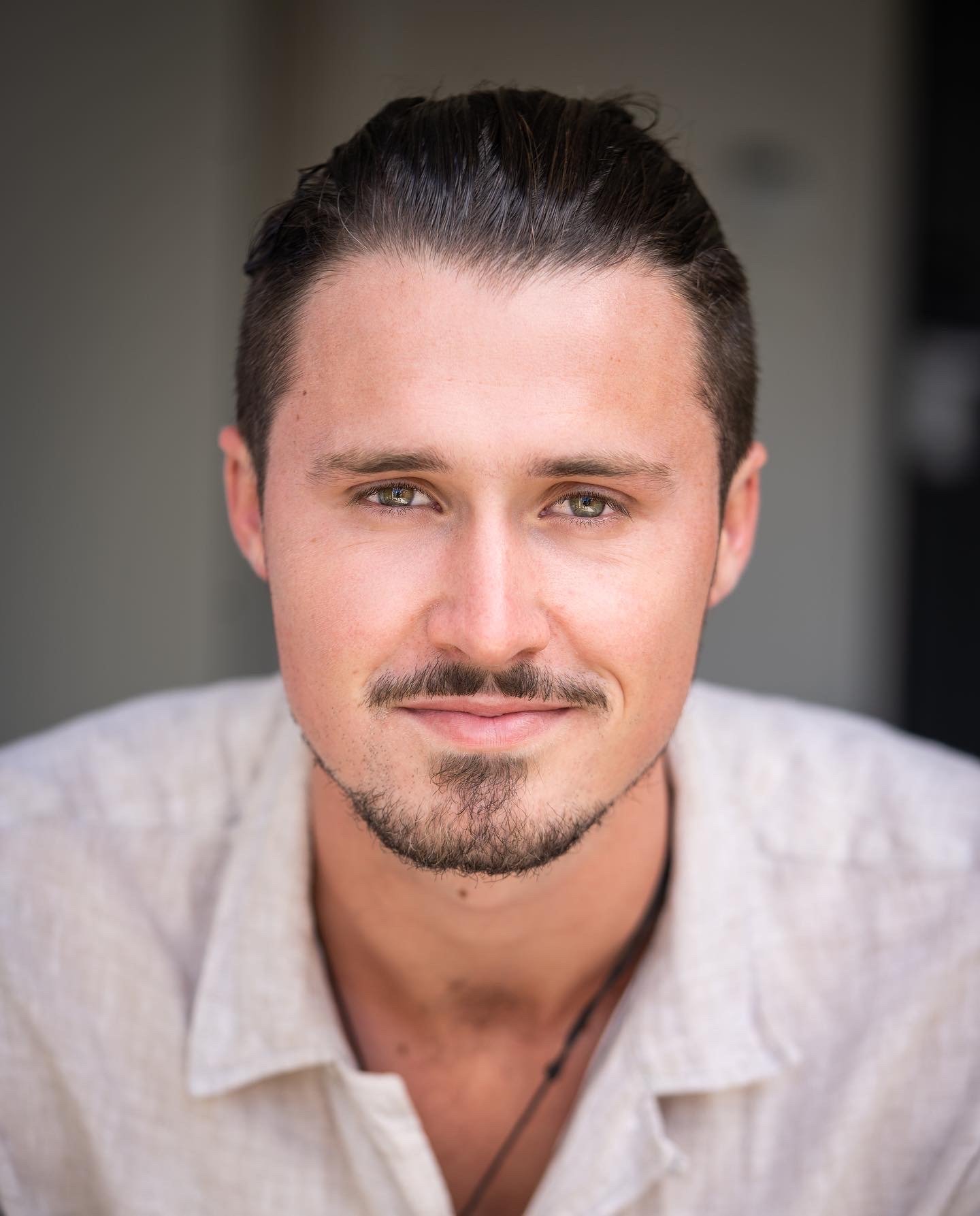Close-up portrait of a smiling man with dark hair, light skin, green eyes, and a goatee, wearing a light-colored shirt.