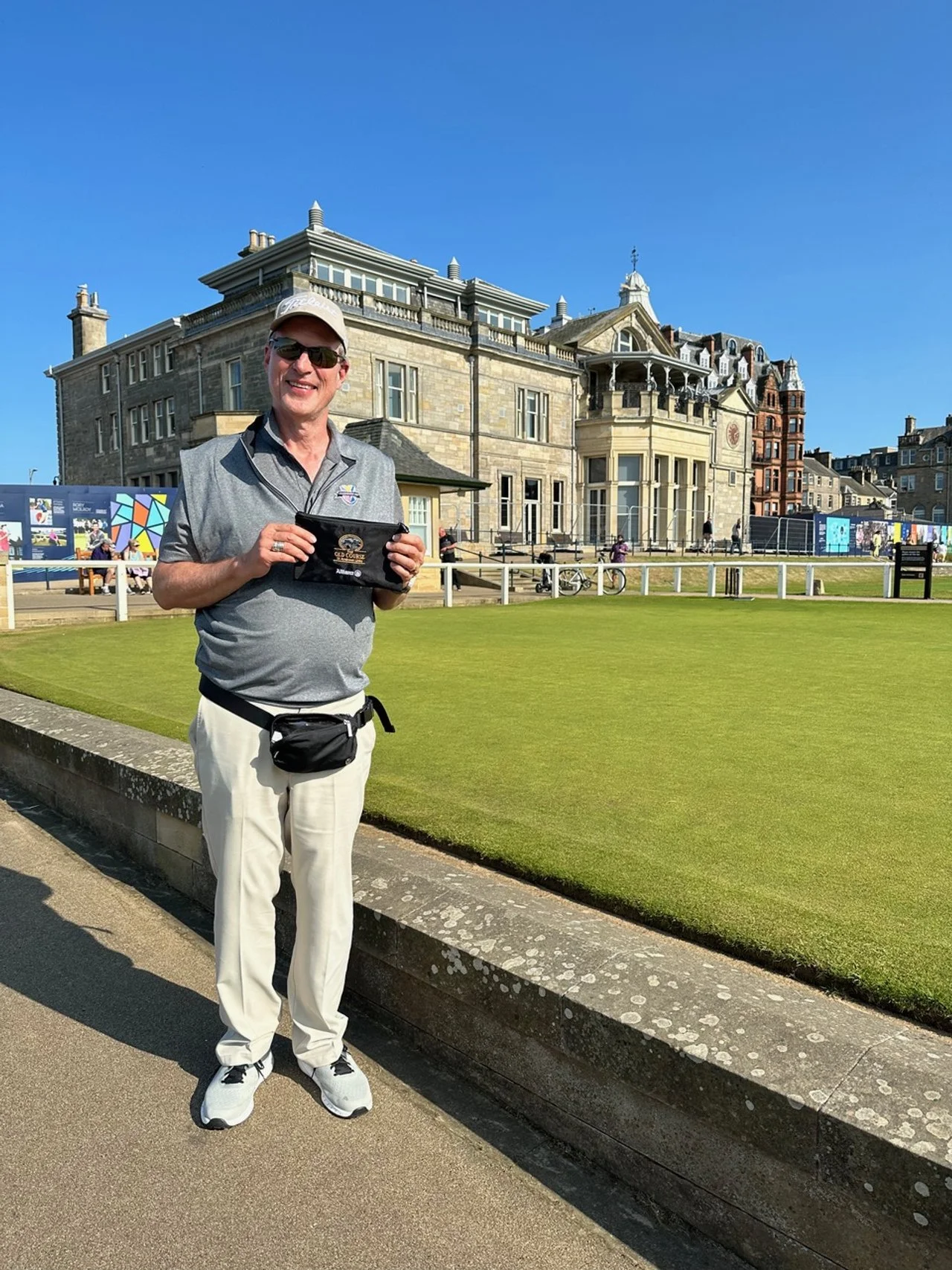 A man standing on a sidewalk near a grass lawn, holding a black tablet, wearing sunglasses, a gray polo shirt, white pants, a beige cap, and a black waist pouch, in front of a historic stone building with large windows and balconies, under a clear blue sky.