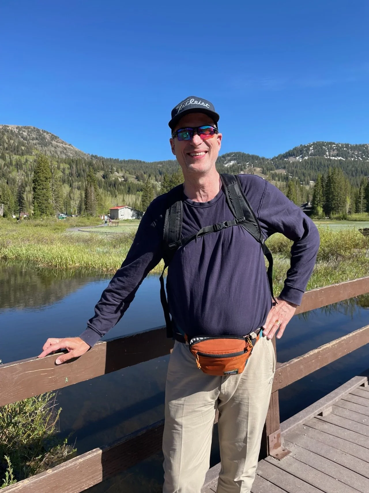 A man wearing sunglasses, a hat, a long sleeve shirt, and a backpack standing on a wooden bridge with a scenic mountain and forest background.
