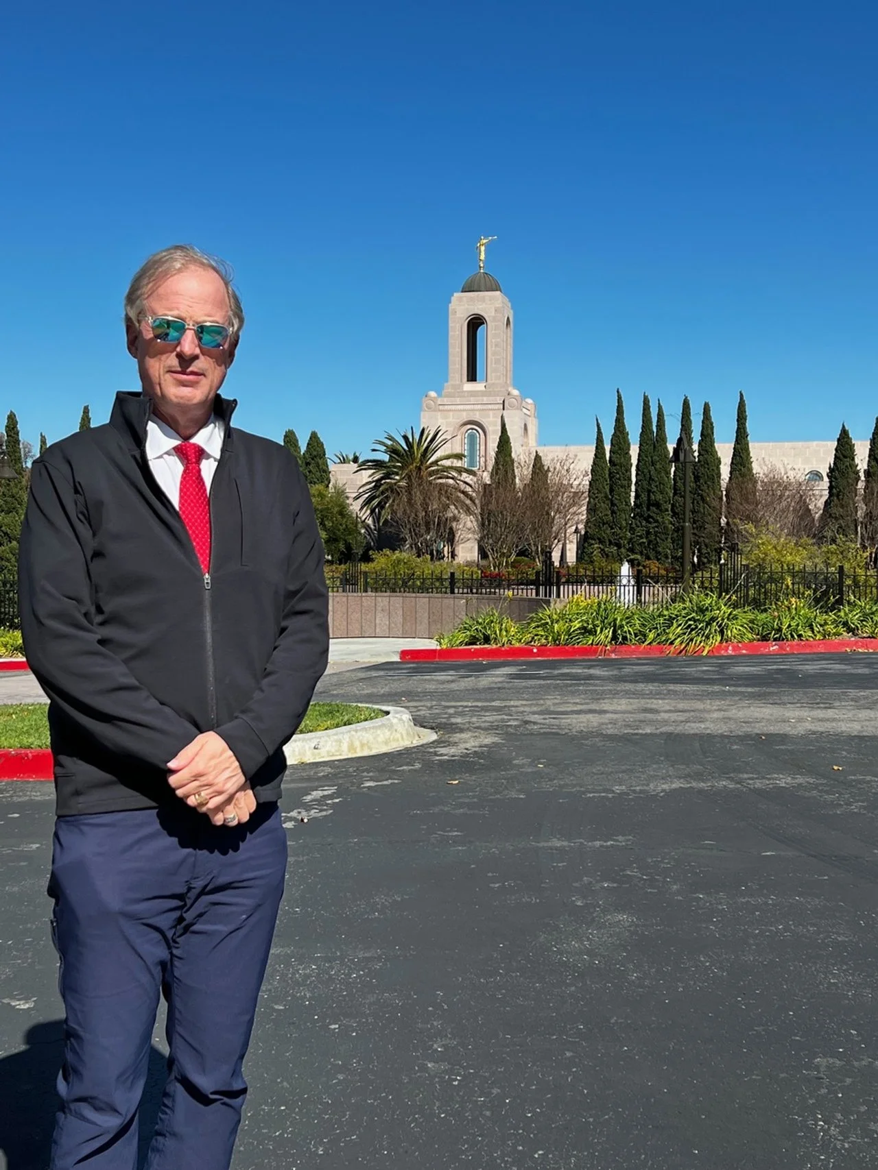 A man wearing sunglasses, a black jacket, a white shirt, and a red tie stands with folded hands on a paved surface. Behind him is a historic church with a tall bell tower topped with a gold weather vane, surrounded by trees and greenery under a clear blue sky.