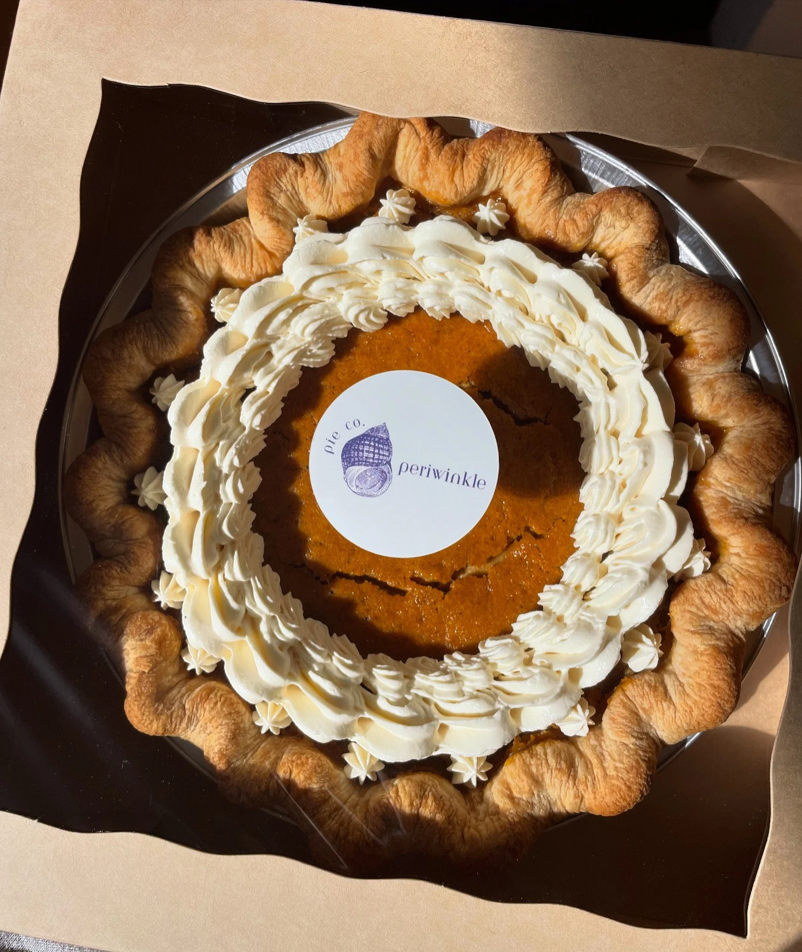 A pumpkin pie decorated with whipped cream, on a black plate with a paper border. The pie has a crust border and a label in the center reading 'Periwinkle Pie Co.'.