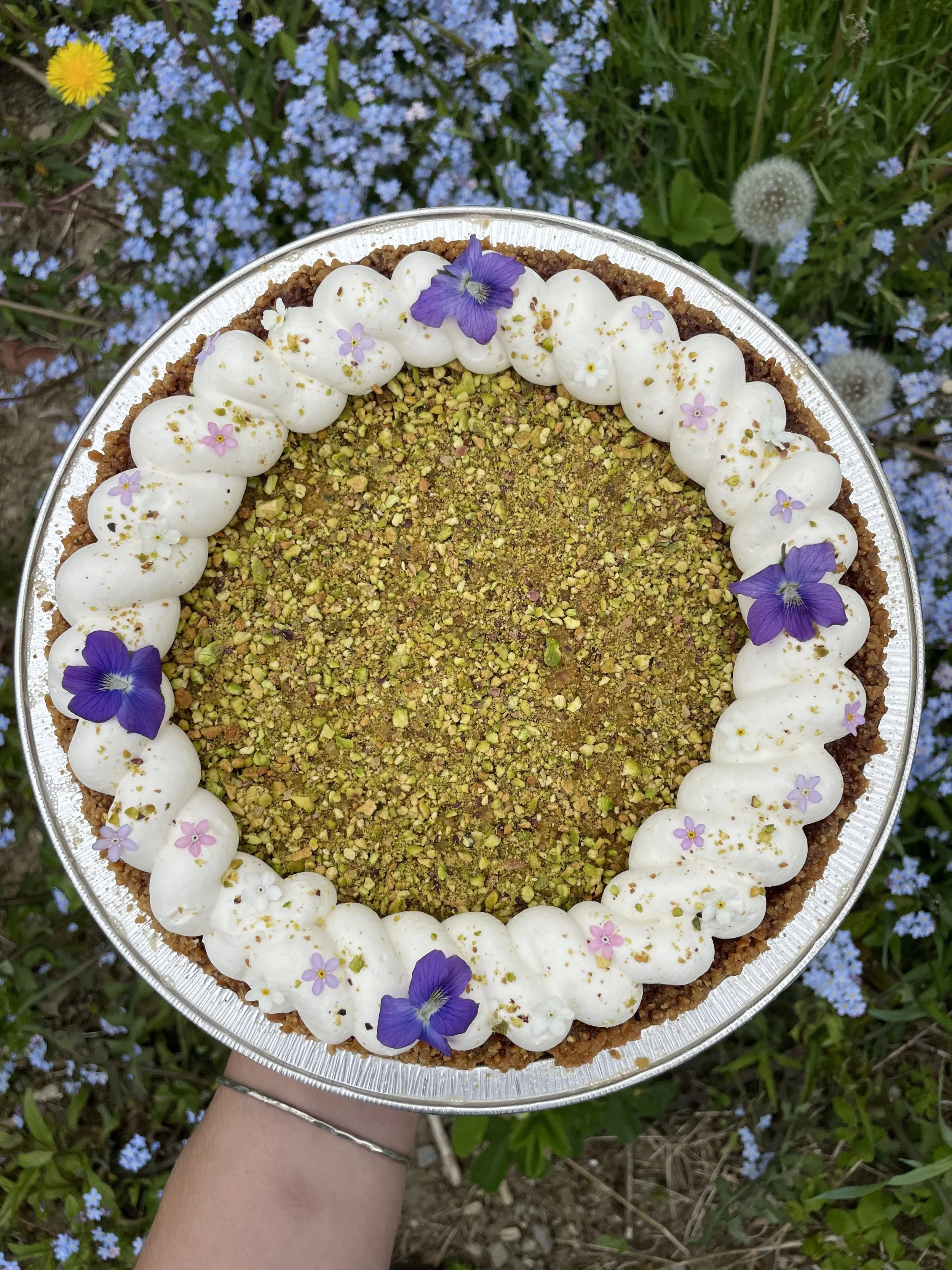 A round cake topped with whipped cream, chopped pistachios, and decorative edible flowers, held above a bed of blue flowers.