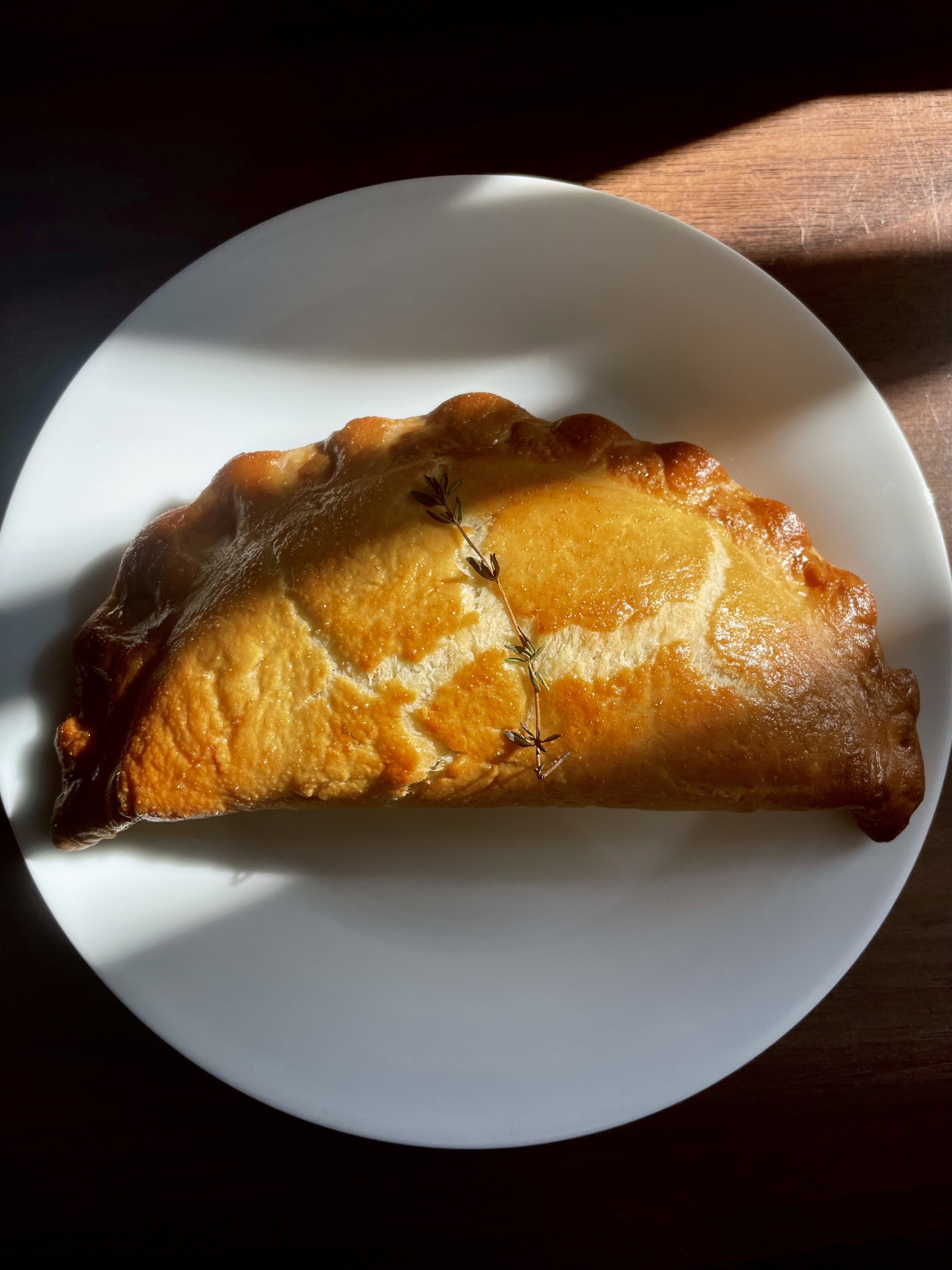 A golden baked calzone with a small sprig of thyme on top, served on a white plate with natural lighting.