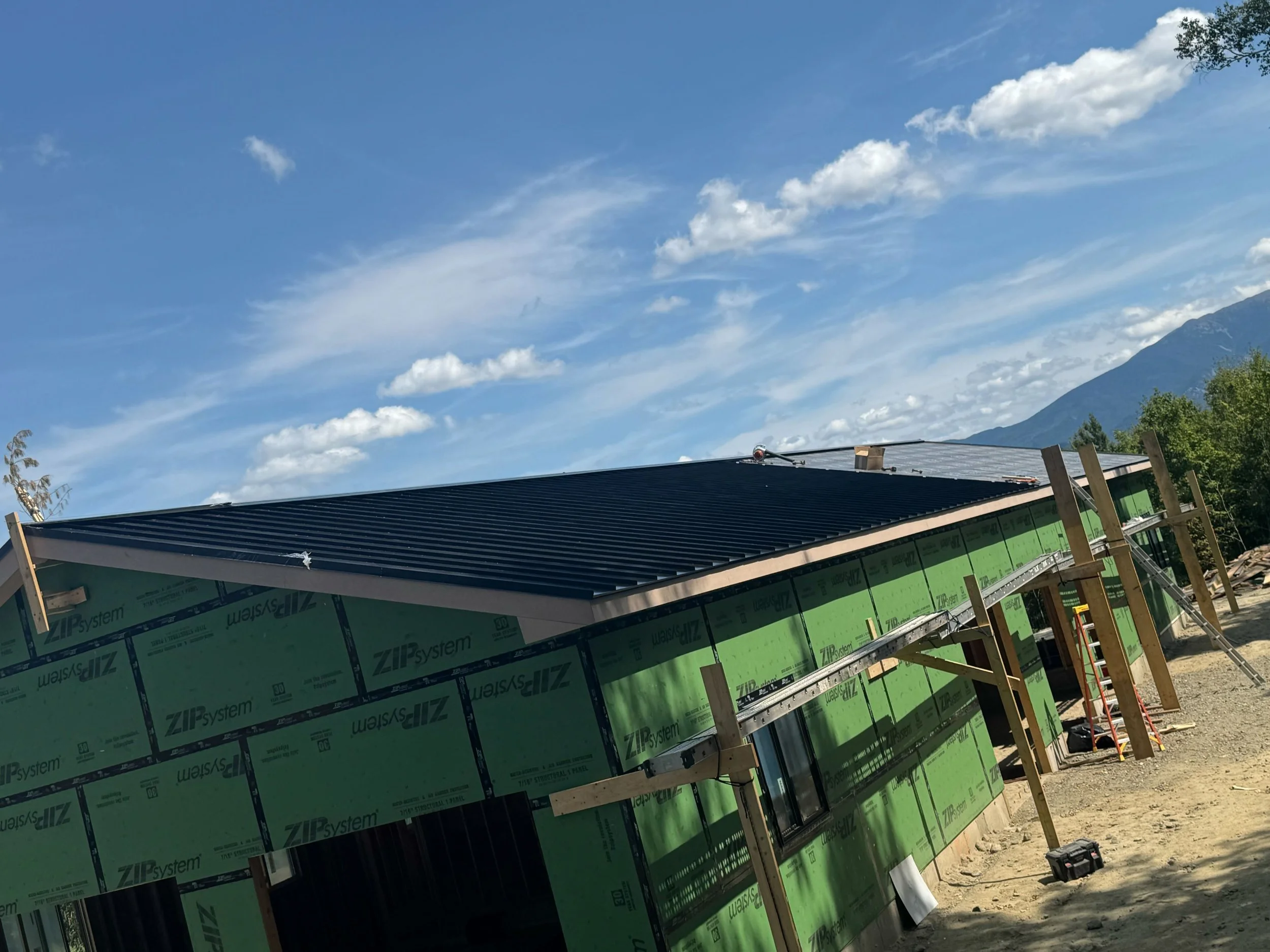 A house under construction with a black metal roof, green building wrap, wooden supports, and various tools, set against a background of blue sky with clouds and distant mountains.