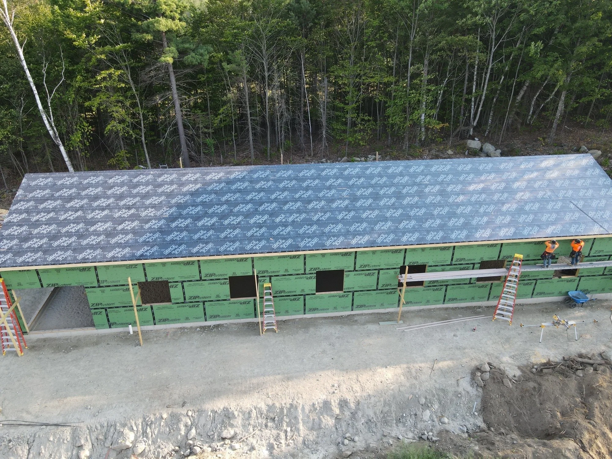 Construction site with a building in progress of roof, workers in safety vests on the roof, and ladders leaning against the structure.