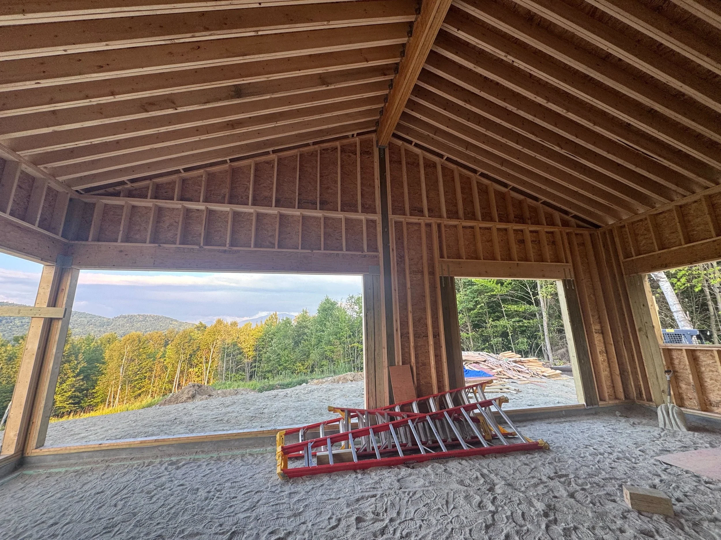 Interior view of a house under construction with exposed wooden framing, large window openings, and a ladder on the unfinished sandy floor.