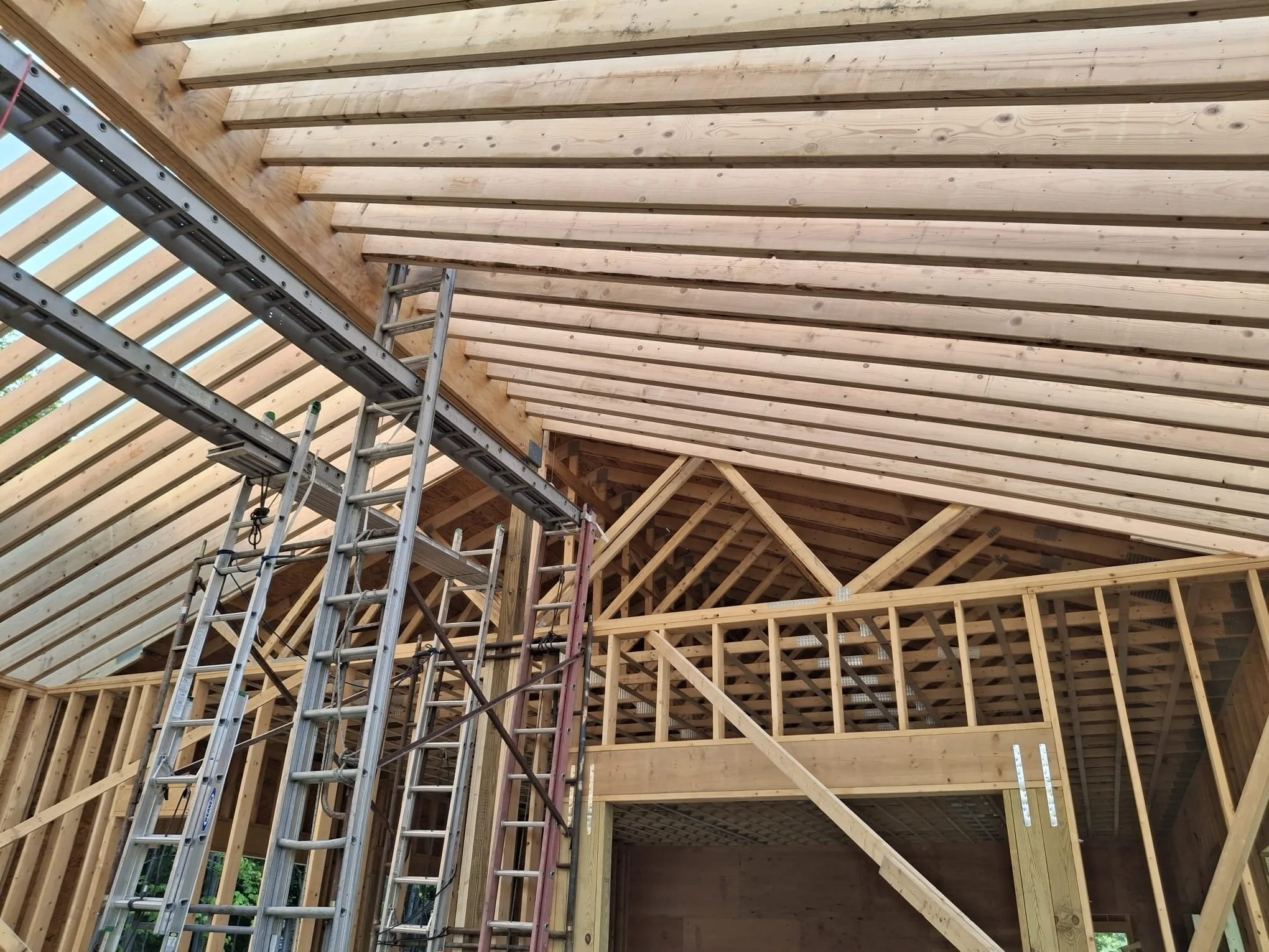 Interior view of a house under construction with exposed wooden framing, scaffolding, and roof trusses.