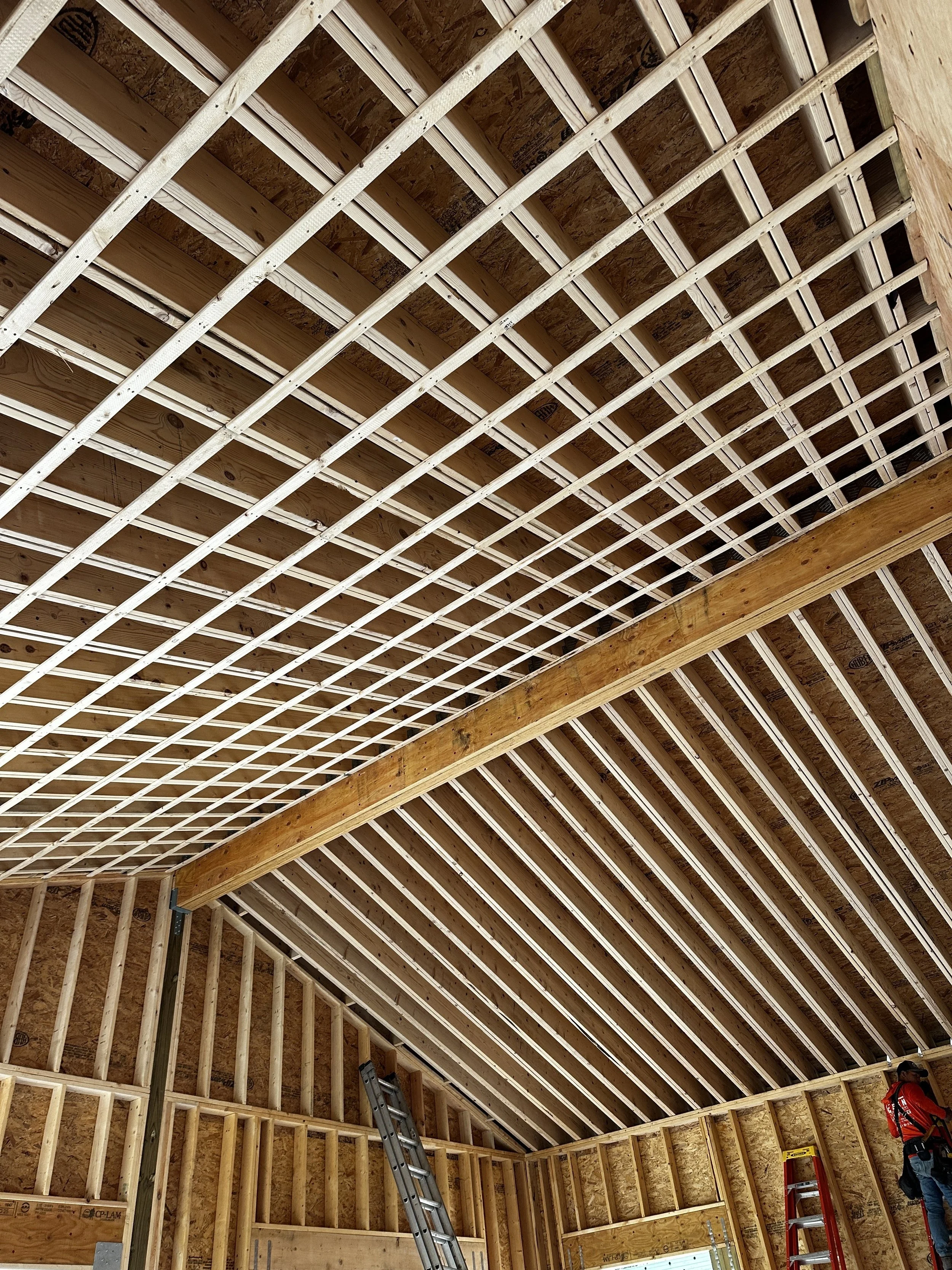 Interior view of a building under construction with wooden framing and a ceiling with crossed beams, construction worker standing on a ladder, and construction tools and materials in the background.