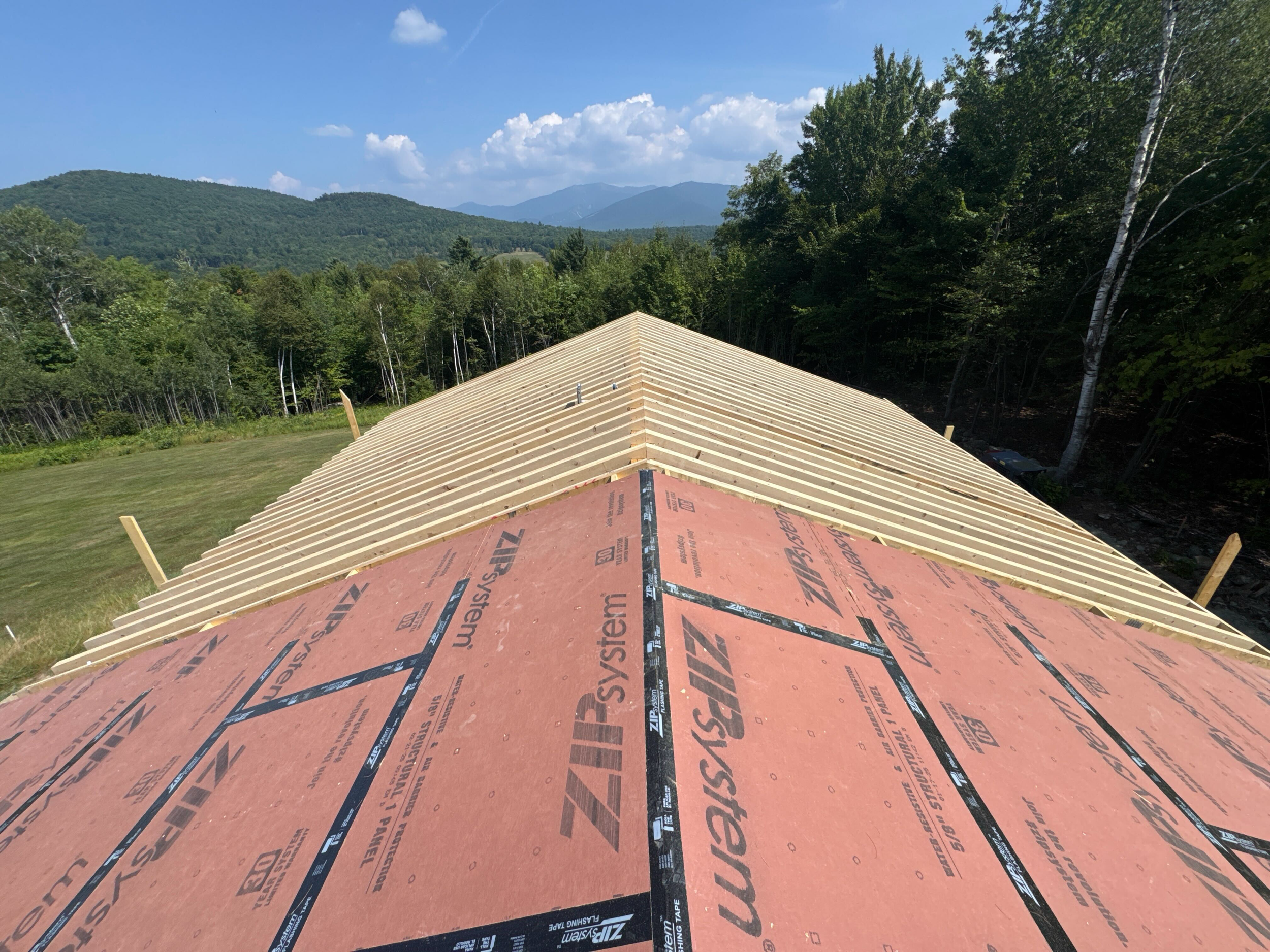 Roof under construction with exposed wooden roof trusses and red underlayment, surrounded by green trees and distant mountains under a partly cloudy sky.