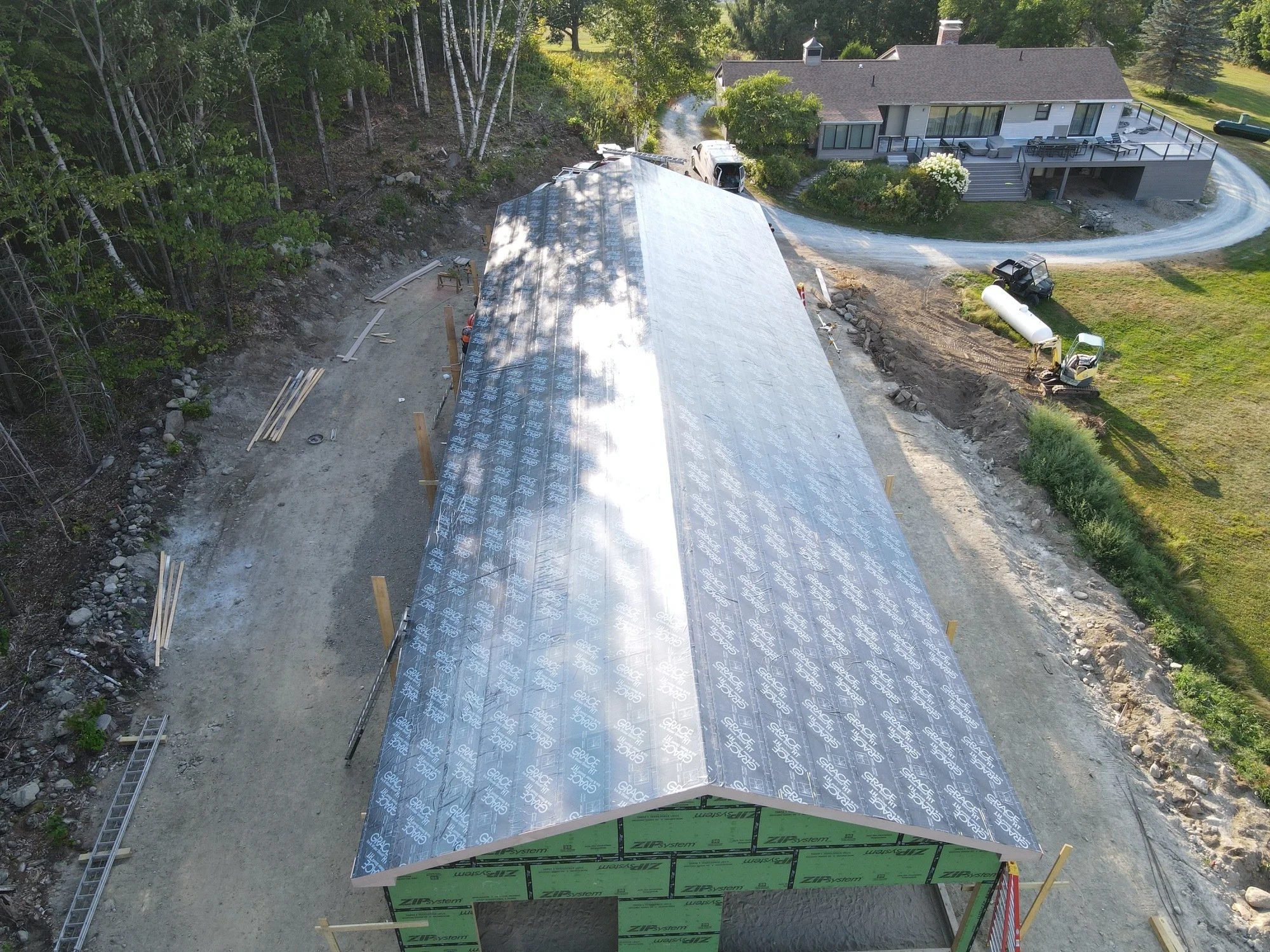 An aerial view of a construction site showing a large building with a metal roof partially covered in reflective weather barrier, surrounded by dirt and construction materials, with a house and trees in the background.