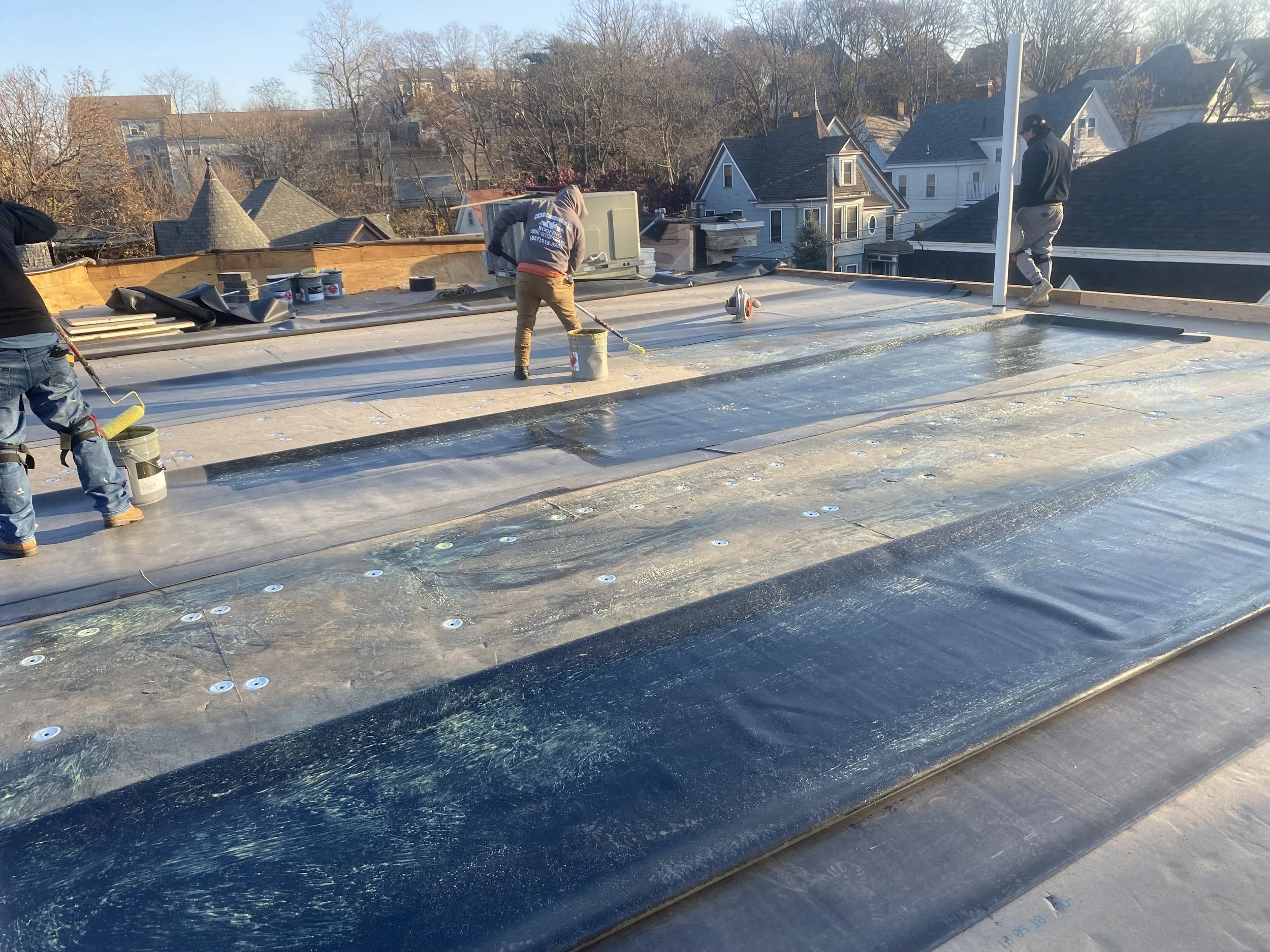 Three workers applying a waterproofing coating on a flat rooftop during daytime, with residential buildings and trees in the background.