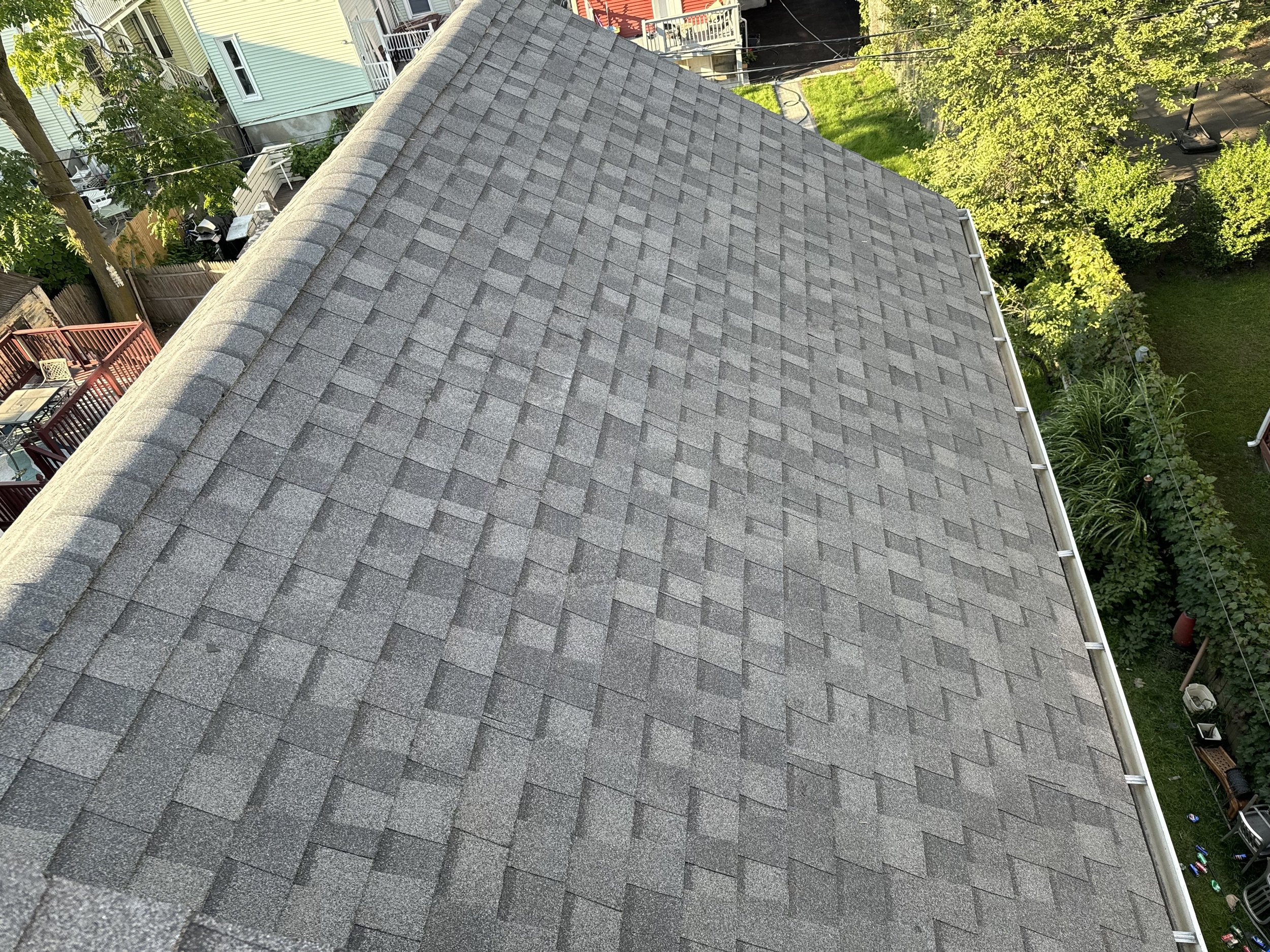 View of a gray shingled roof of a building, with trees and other houses in the background.