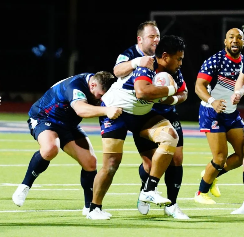 American rugby players in blue and stars and stripes uniforms celebrating during a game, with one player holding a rugby ball and others supporting him.