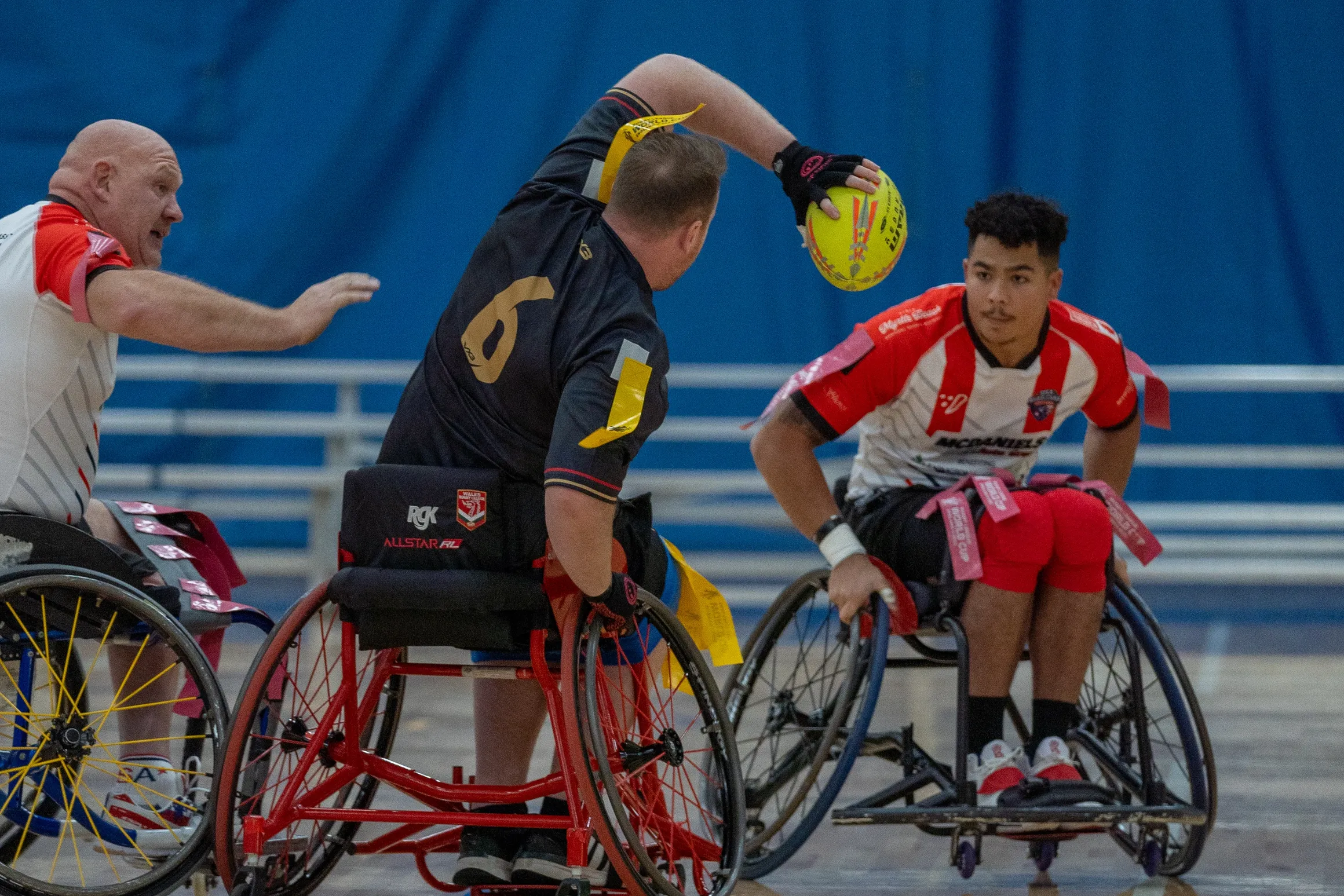 Wheelchair rugby players and a referee on an indoor court, one player holding a yellow ball, and others in sports gear.
