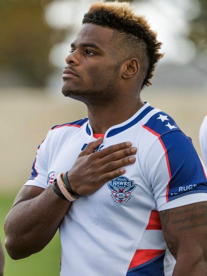 A man with short, styled hair and a beard, wearing a USA Rugby team jersey with the USA Rugby Hawks logo, placing his right hand over his heart during the national anthem.