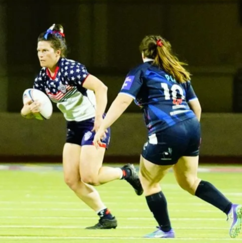 Two female rugby players on a field, one in a star-spangled jersey holding a rugby ball, the other in a blue jersey approaching defensively.