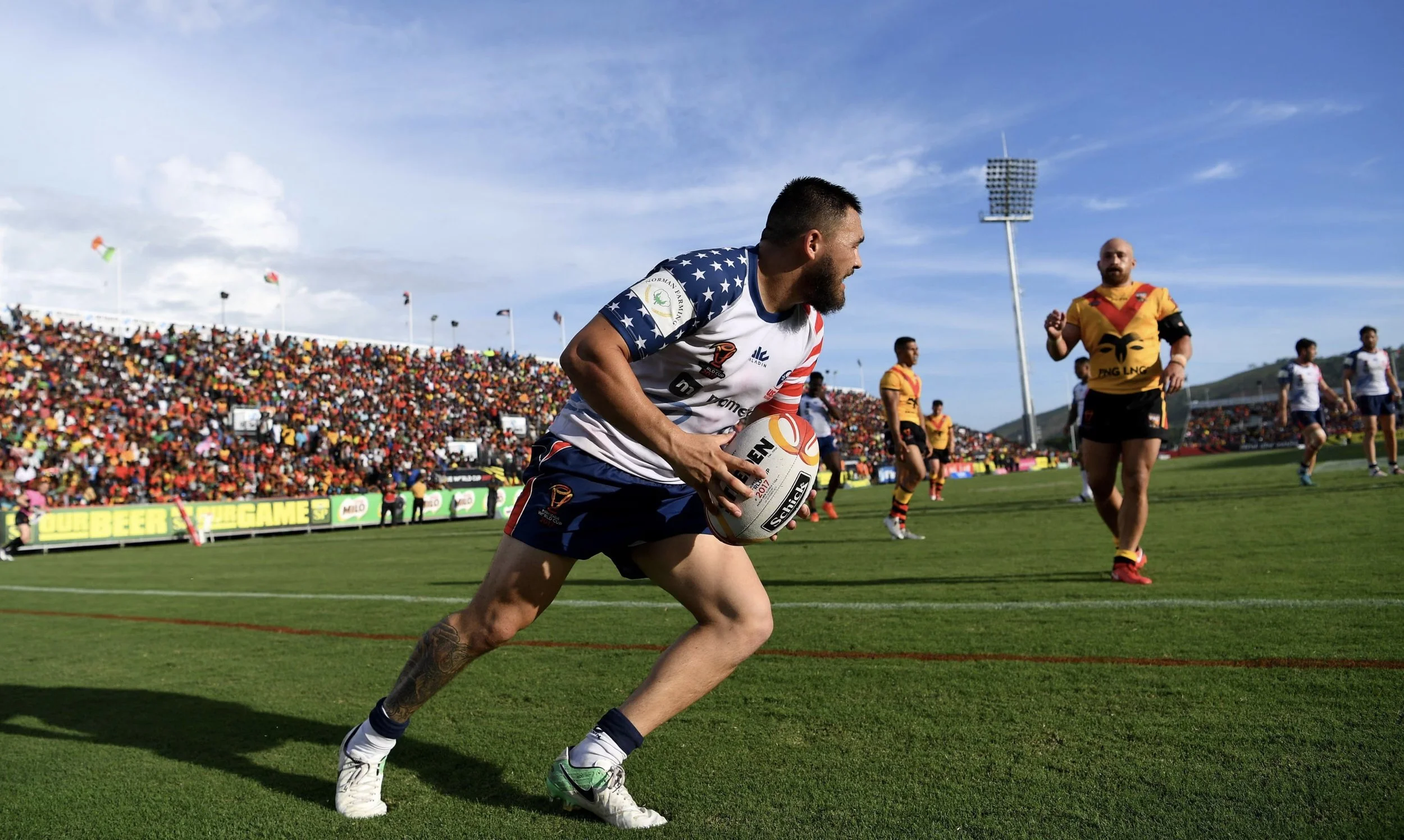 Rugby player in a white jersey with stars on the sleeves holding a rugby ball and running on a green field, with spectators in the stands and other players in yellow and black jerseys in the background under a clear sky.