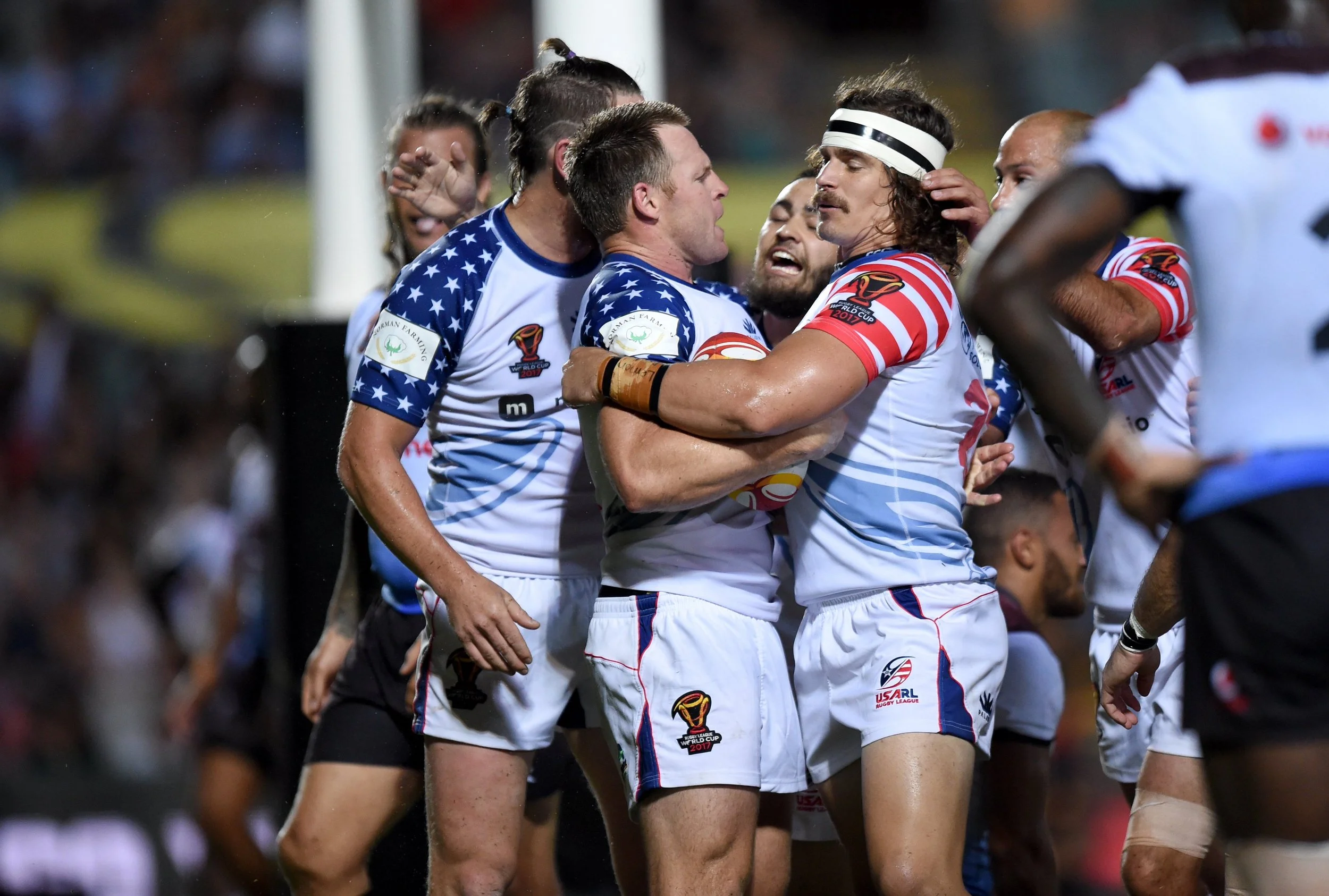 Rugby players from two teams celebrating on the field after a match; one team is wearing blue and white uniforms with a World Cup logo, and the other team is wearing red, white, and blue uniforms. The players are hugging and showing excitement.