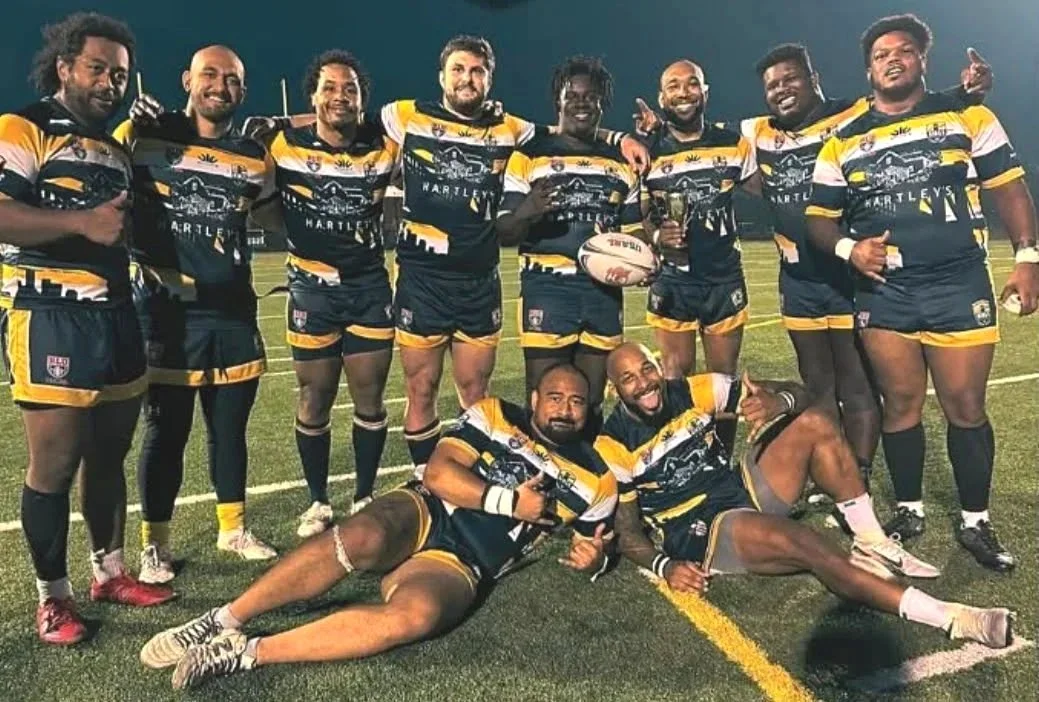 A group of ten rugby players in black and yellow uniforms on a field, with two players lying in front and the others standing behind, holding a rugby ball and celebrating.